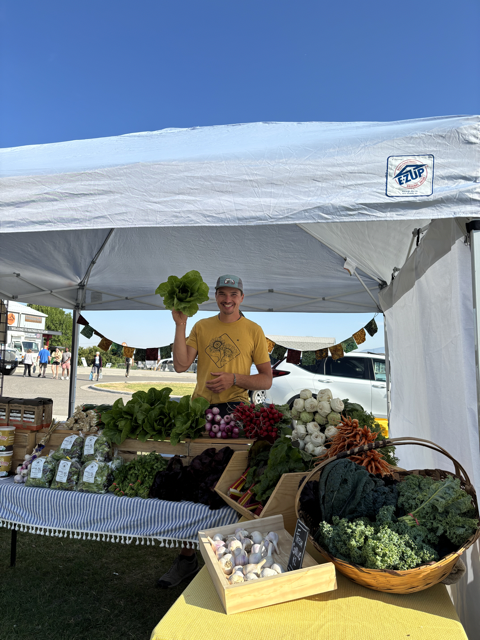 Farmer at outdoor market stand selling fresh vegetables and greens under a white canopy, with a person holding a head of lettuce and smiling.