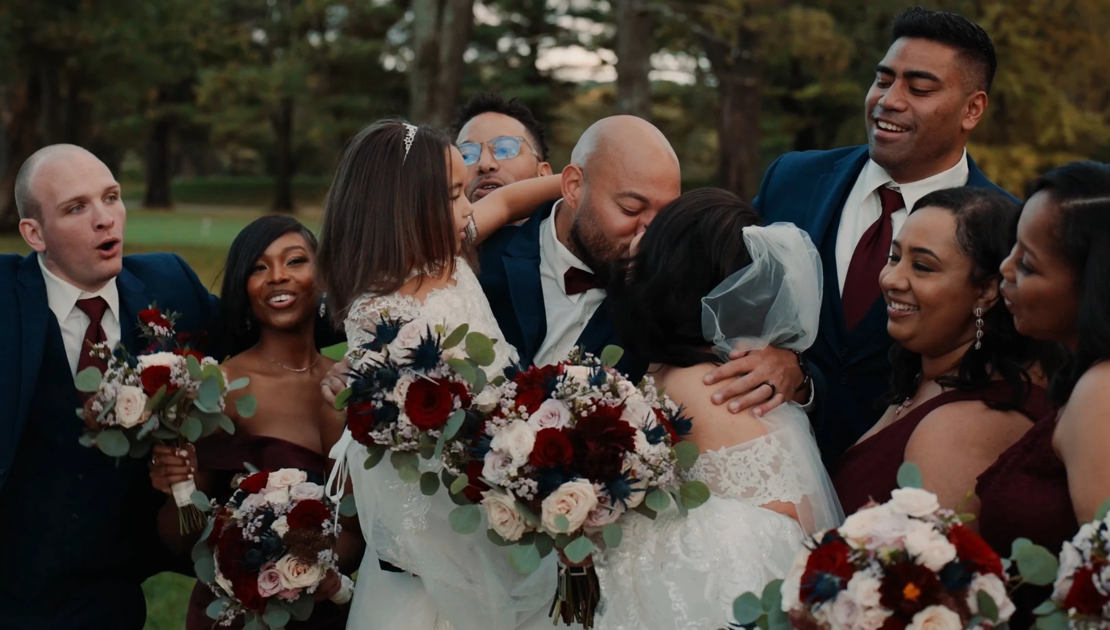 Group of people at a wedding, embracing, smiling, and holding bouquets of flowers outdoors in a park with trees in the background.