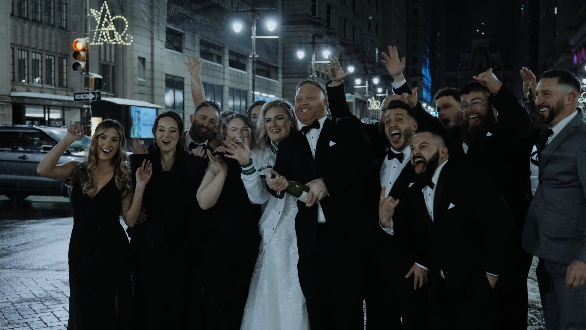Group of people celebrating outdoors at night, dressed in formal attire with some in tuxedos, holding champagne bottles, smiling and making joyful gestures.