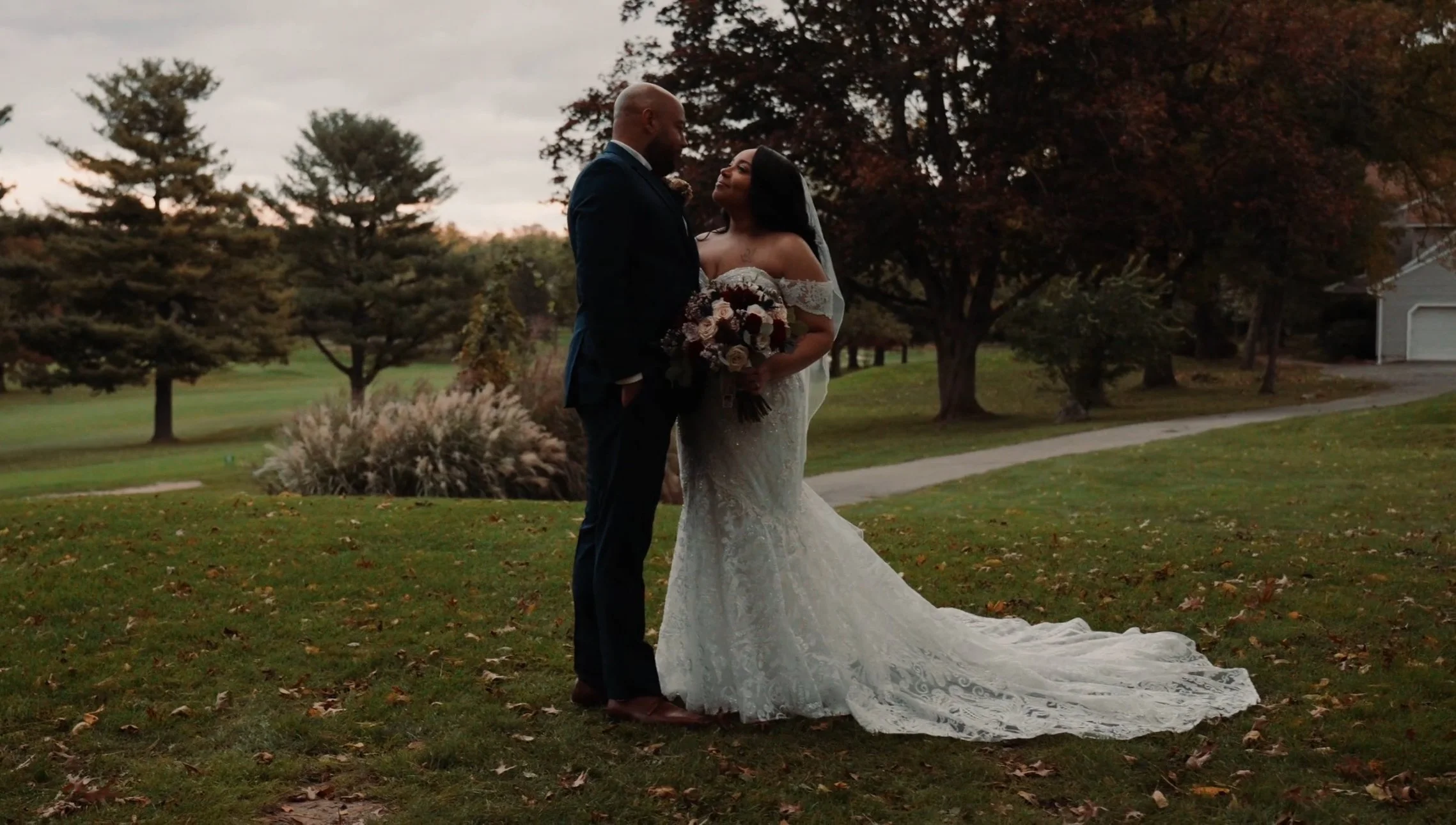 A bride and groom standing close in a park during sunset, with trees and a house in the background. The bride is holding a bouquet and wearing a white lace wedding gown, while the groom is in a dark suit.