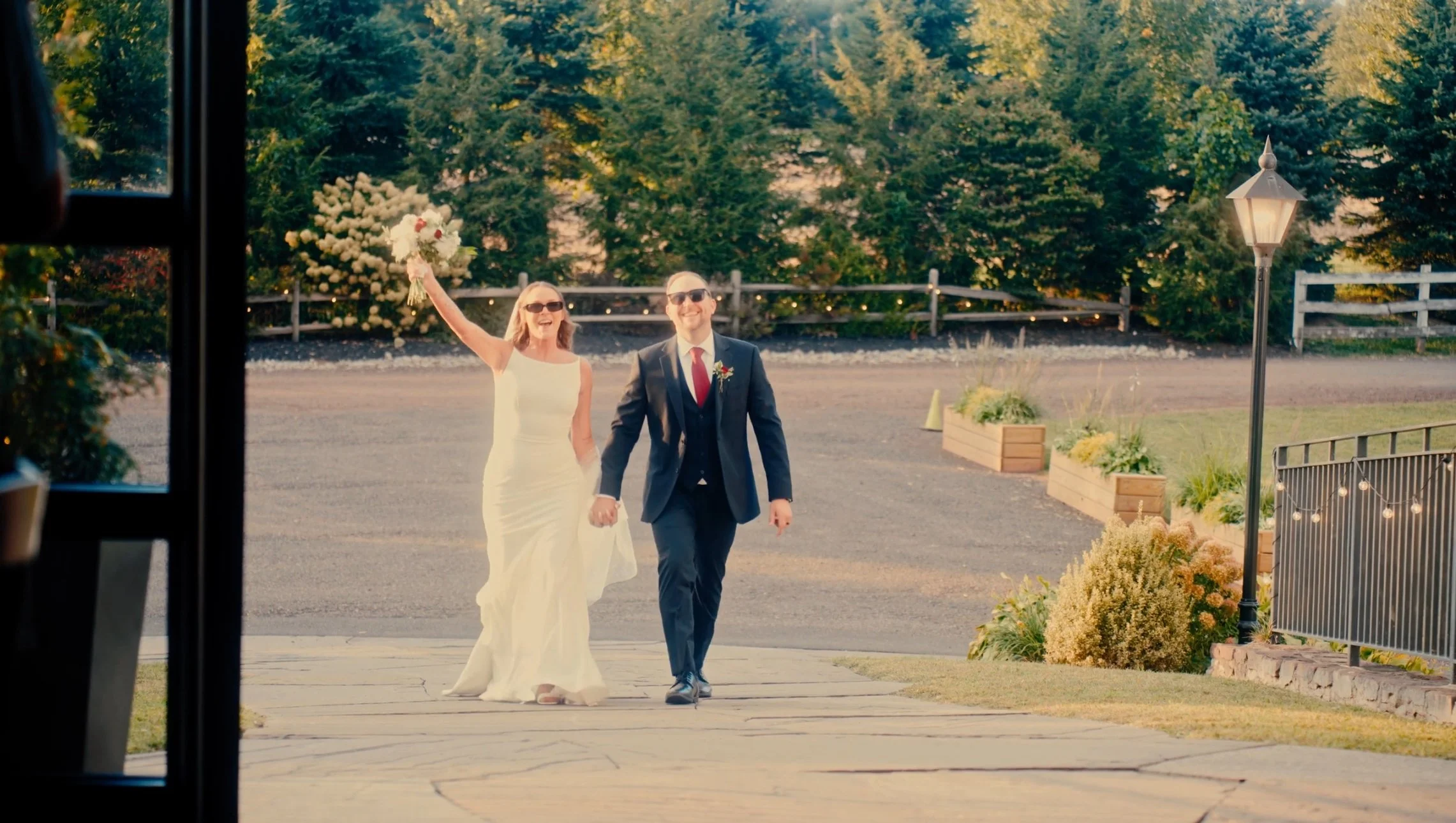 A newlywed couple walking hand in hand outdoors, with the bride holding a bouquet of flowers and raising her arm joyful, and the groom smiling in a suit and sunglasses, in a garden setting during daytime.