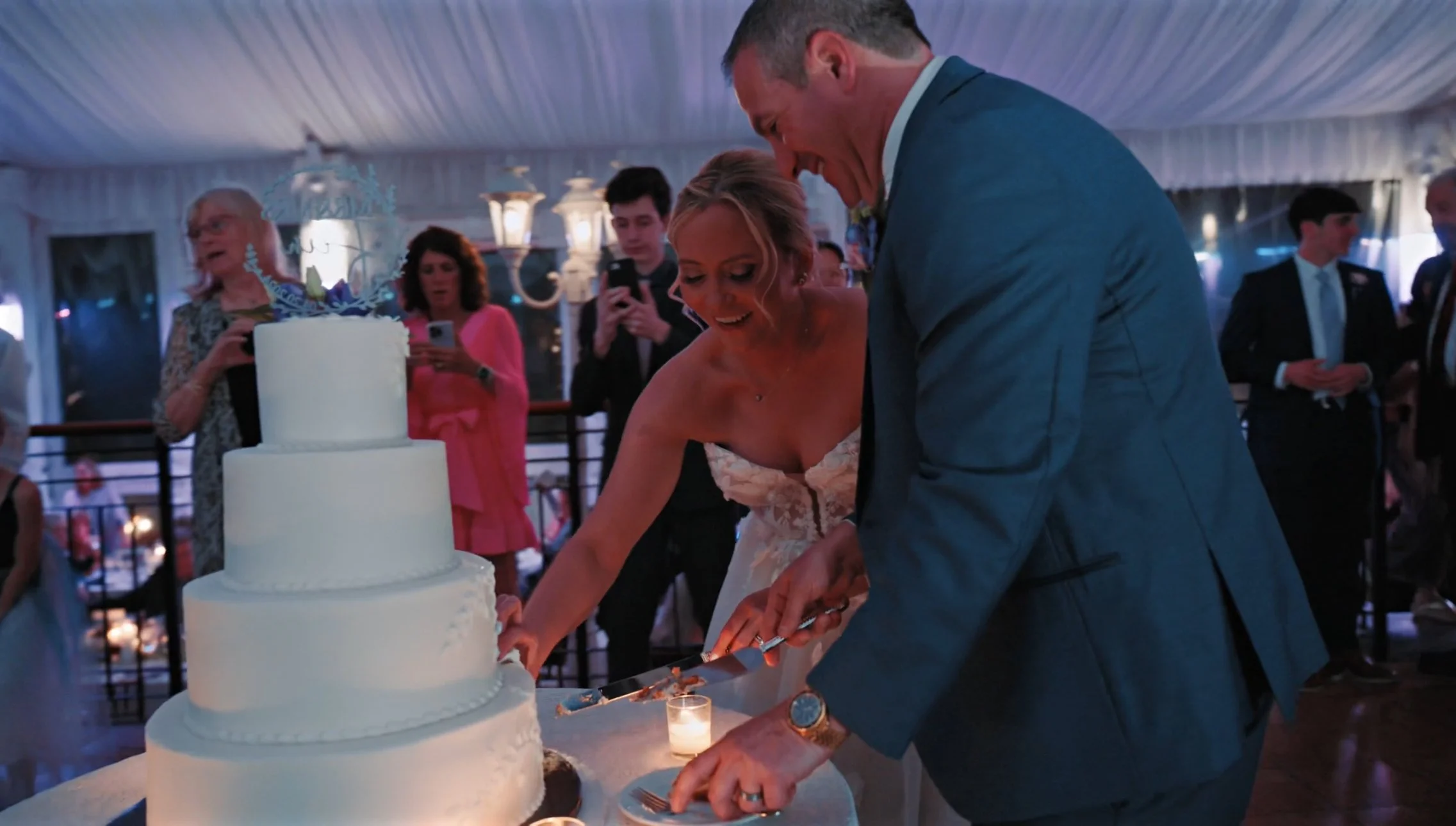 A bride and groom cut their wedding cake together at a celebration. The wedding cake is a four-tiered white cake with a decorative topper, and the couple is smiling and laughing. Guests are in the background, some taking photos.