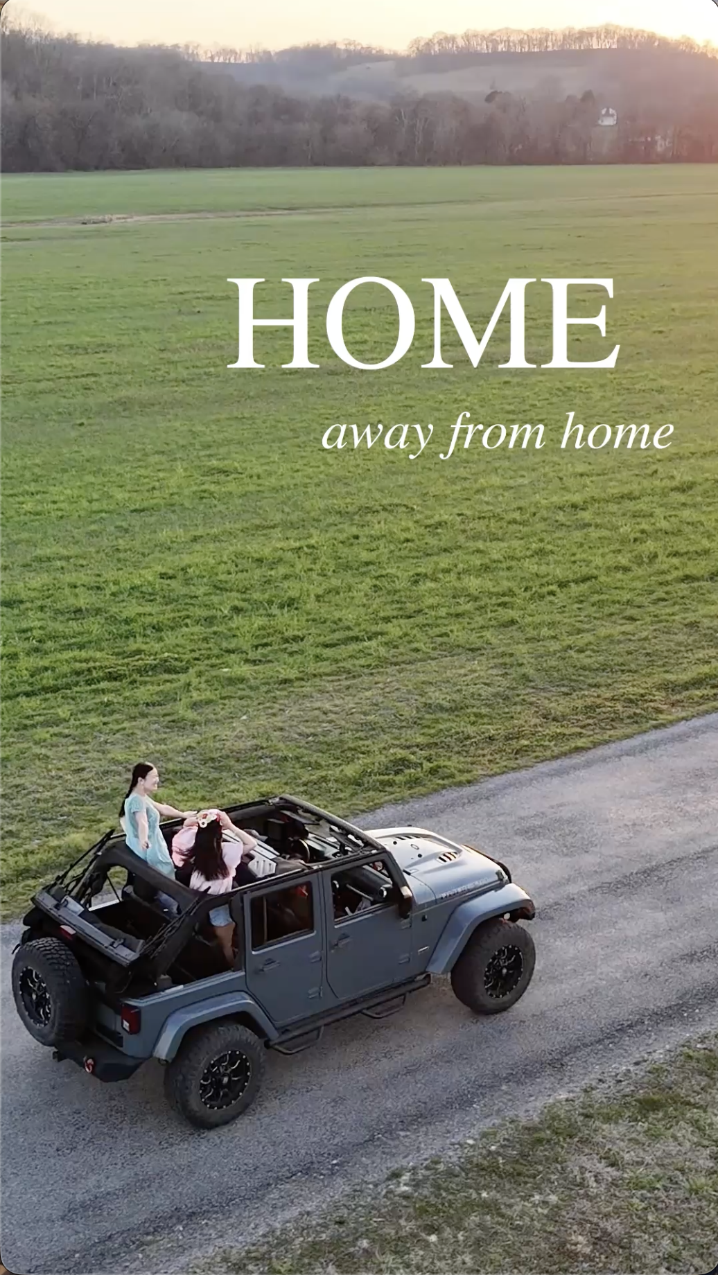 An aerial view of a blue Jeep driving on a gravel road beside a large green field in Pulaski Tennessee. The scene is outdoors during sunset, with a rural landscape in the background and text overlay saying "HOME away from home."