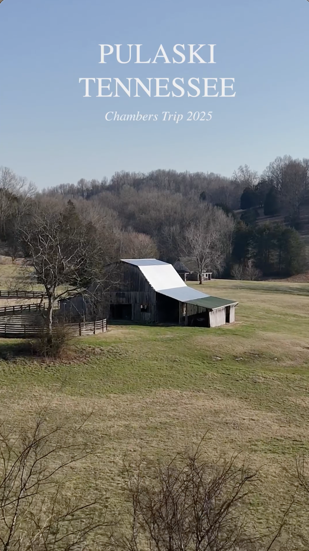 A rural landscape featuring a wooden barn with a white and green roof, surrounded by open grassy fields and leafless trees, with a backdrop of forested hills under a clear sky. Pulaski Tennessee
