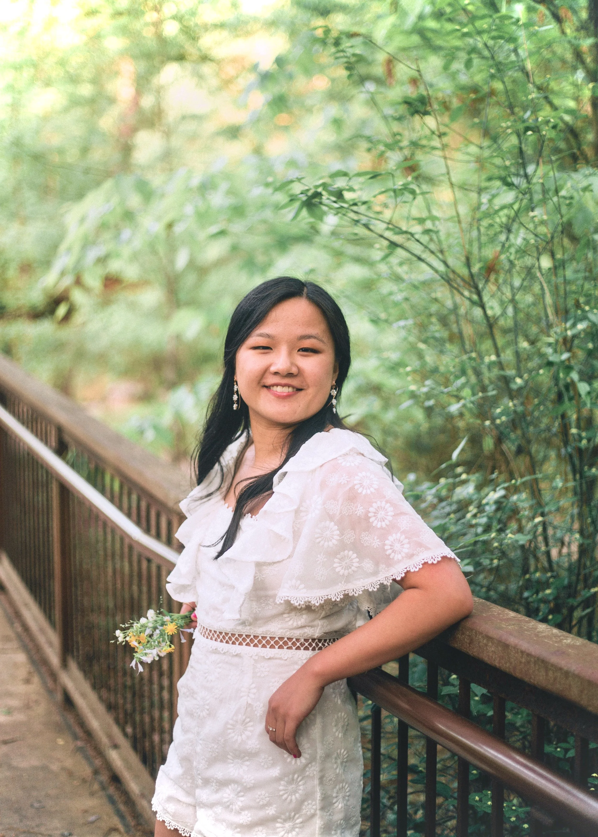 A young woman smiling outdoors, standing by a brown metal railing, surrounded by green trees and foliage, wearing a white dress with floral embroidery and earrings. Lily Chambers graphic design