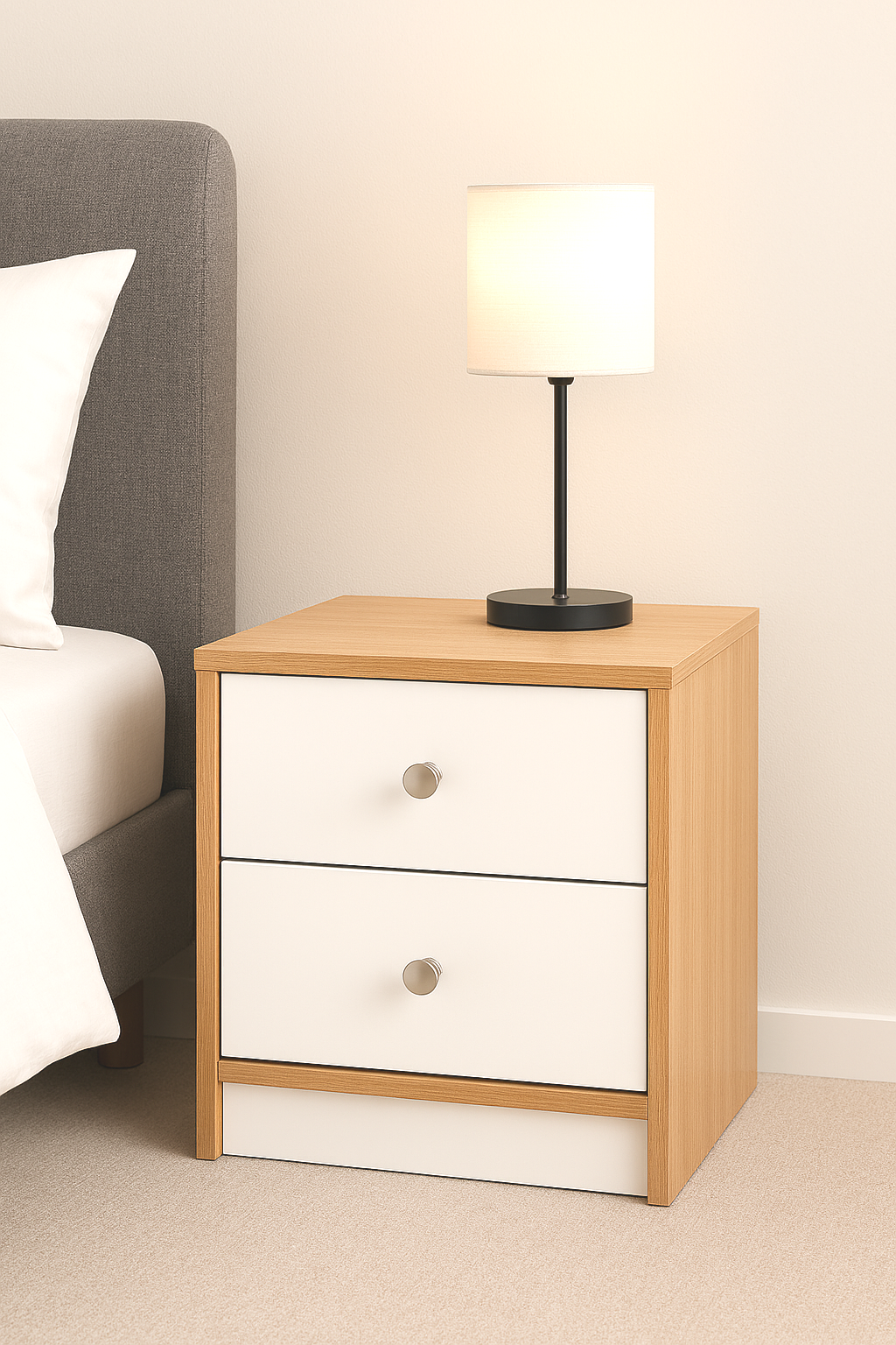 Ein Nachttisch mit zwei weißen Schubladen aus Holz, auf dem eine moderne Lampe mit schwarzem Sockel steht, neben einem Bett mit grauem Kopfteil.