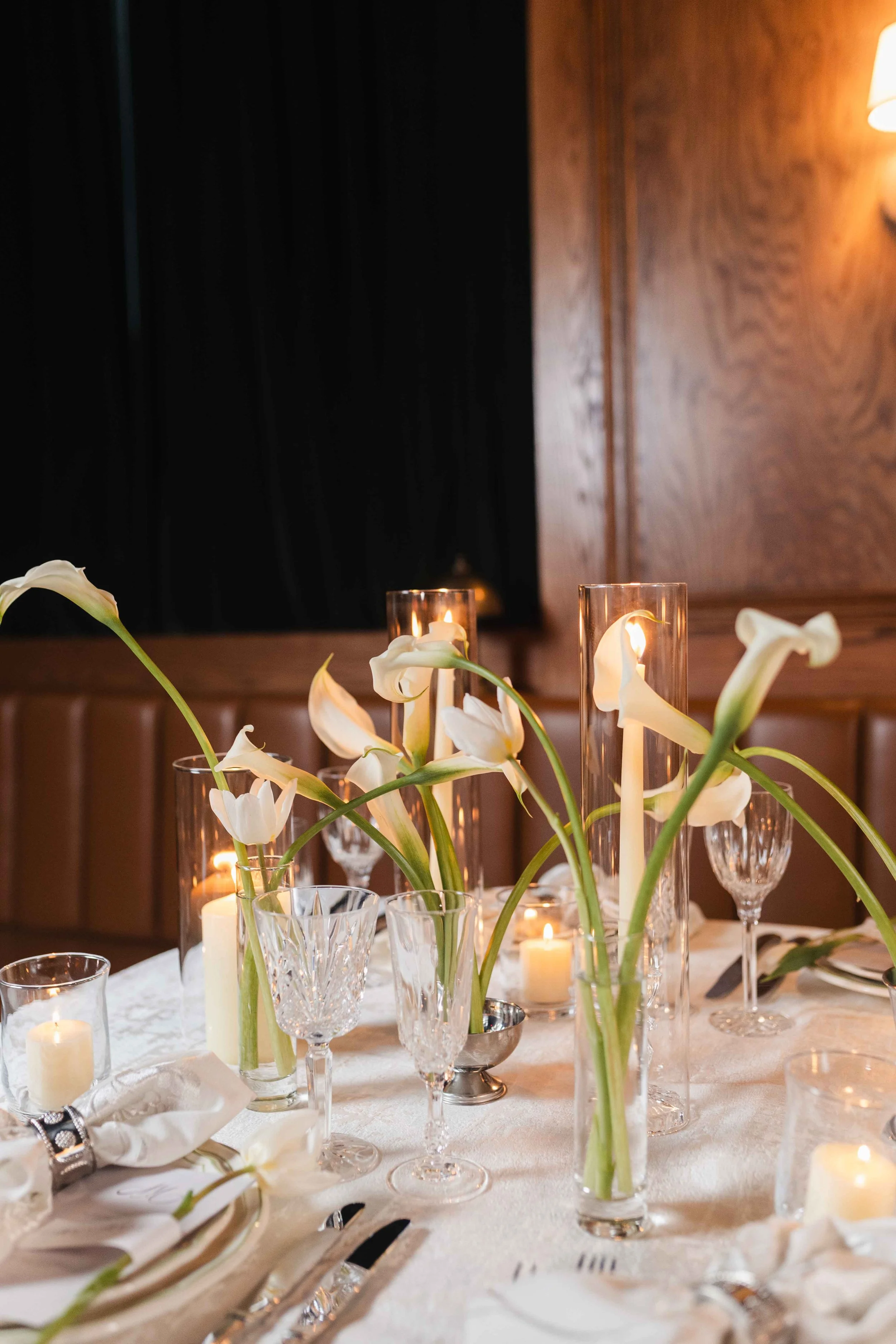 Elegant dinner table centerpiece with white calla lilies in tall glass vases and candles in various holders, set on a white tablecloth.