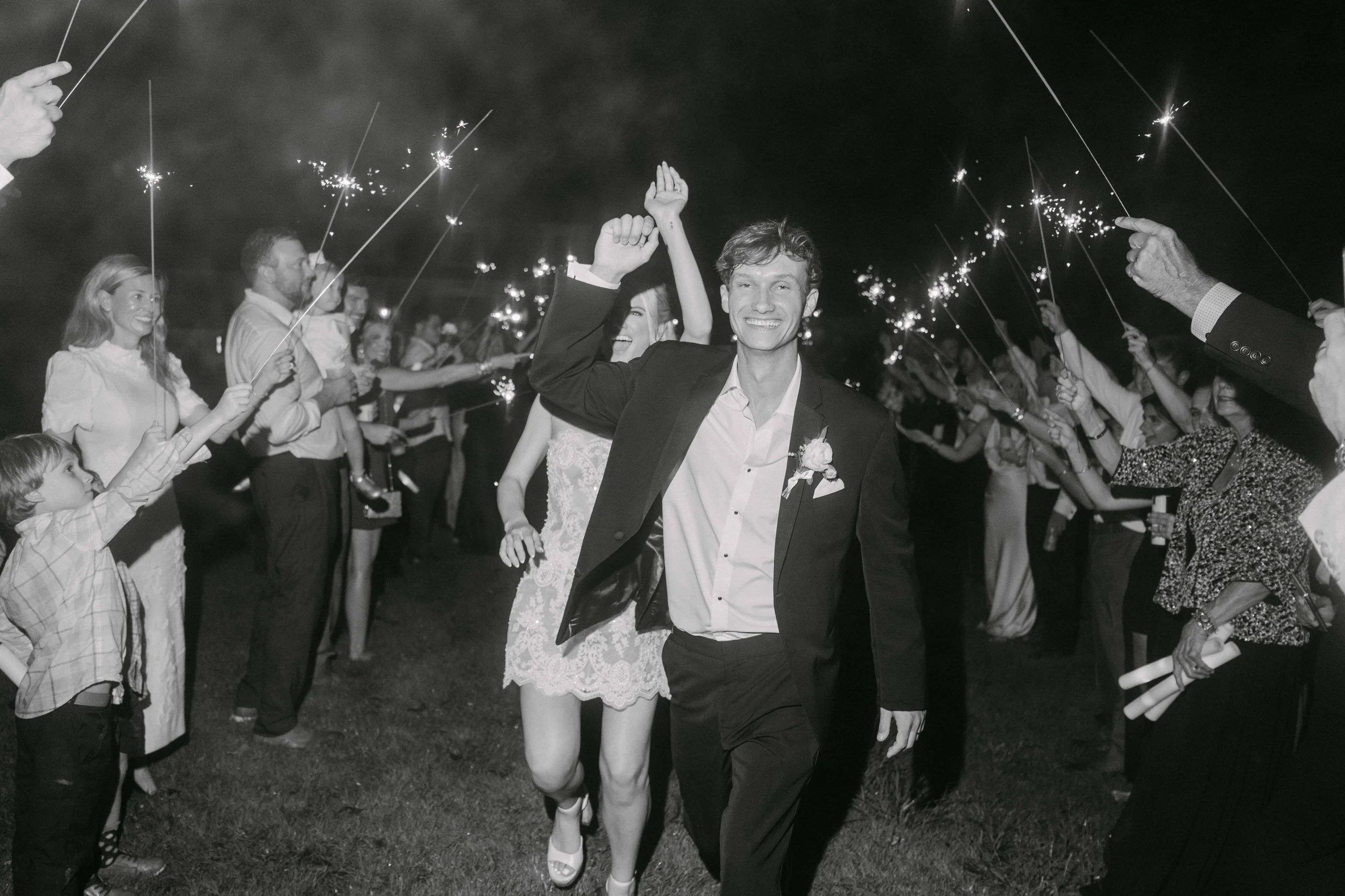 A group of people at a celebration, some holding sparklers, with a smiling man and woman in the center, dressed in formal attire at night. Wedding reception exit.