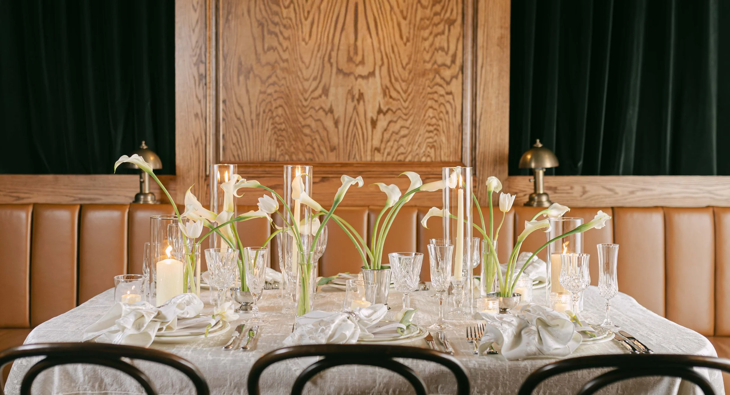 Elegant dining table decorated with white calla lilies in tall vases, surrounded by candles and glassware, in a cozy room with wooden walls and leather seating.