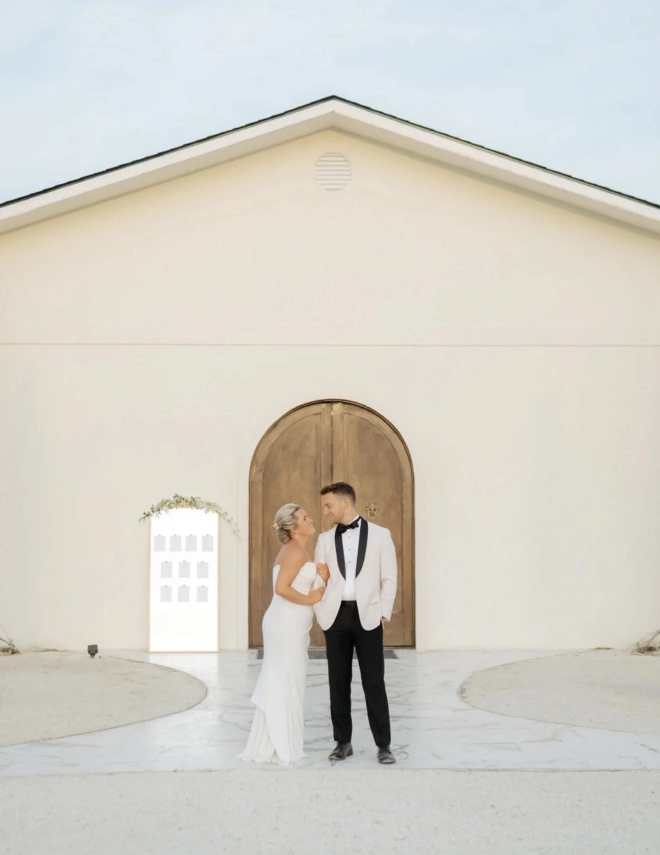 A bride and groom stand together in front of a large wooden archway outside of a white building. The bride is wearing a white wedding dress, and the groom is dressed in a black tuxedo with a white jacket.