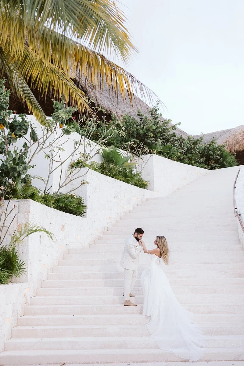 A bride and groom standing on white outdoor stairs, with tropical greenery and thatched roofs in the background, during a wedding celebration.