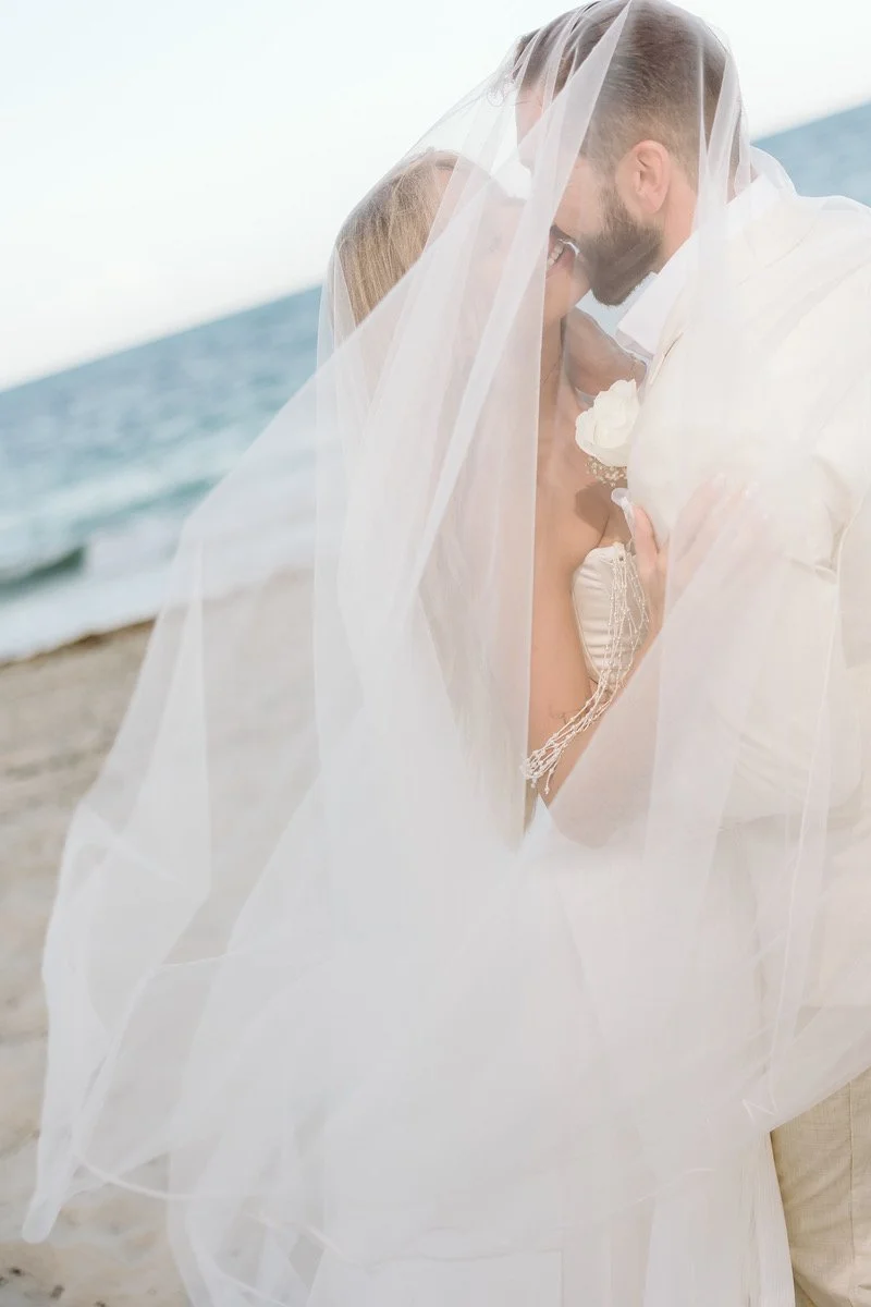 A bride and groom sharing a kiss on the beach under the bride's veil.