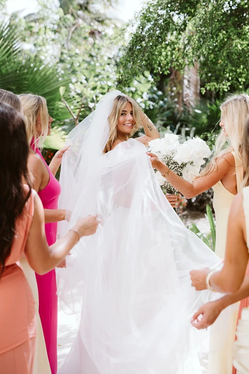A bride smiling and wearing a white wedding dress and veil, holding a bouquet of white flowers, surrounded by several women in colorful dresses in an outdoor garden setting.