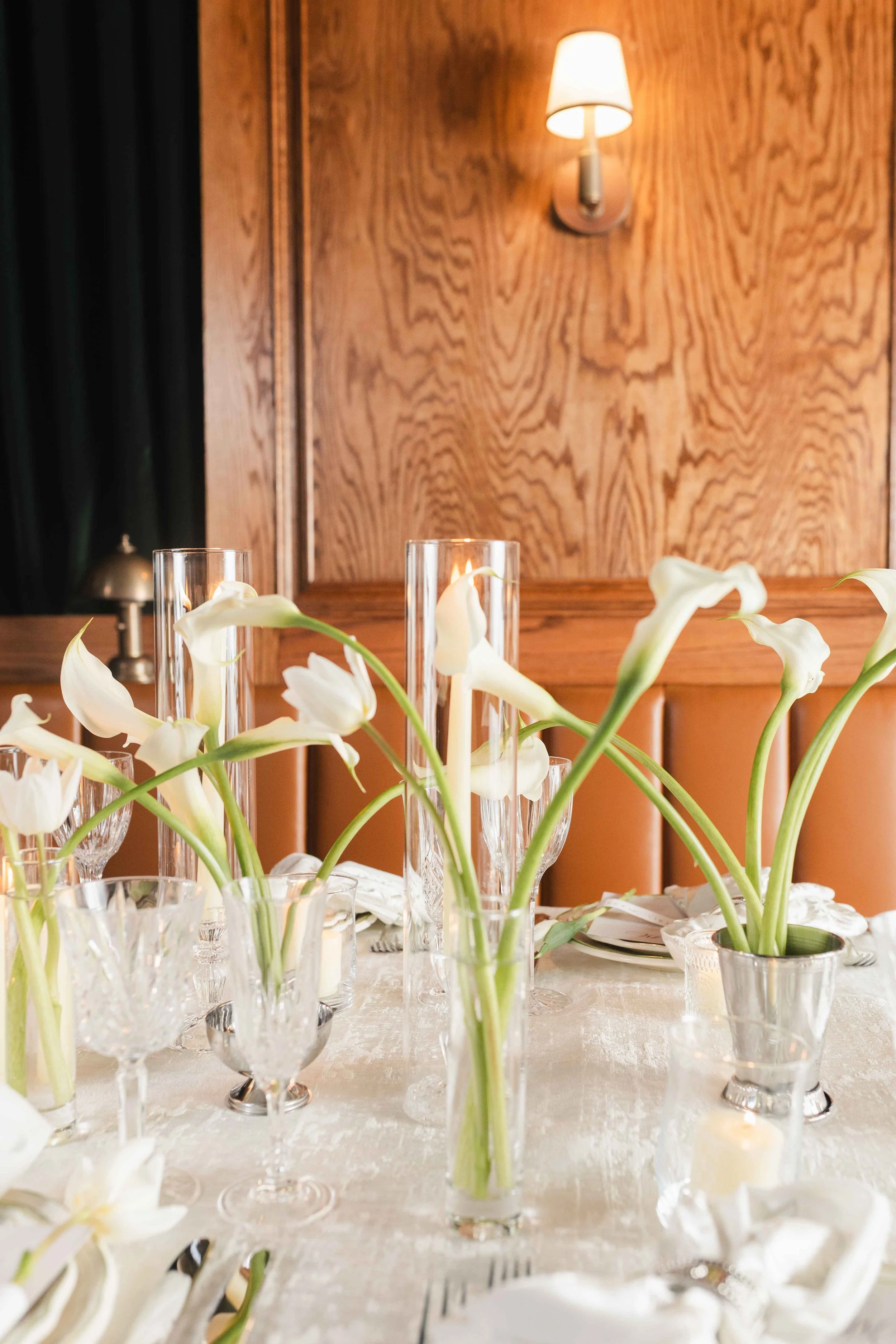 A table decorated with white calla lilies in glass vases, clear glass candle holders, and silver and crystal tableware set on a white tablecloth, with a wooden wall and a wall-mounted lamp in the background.