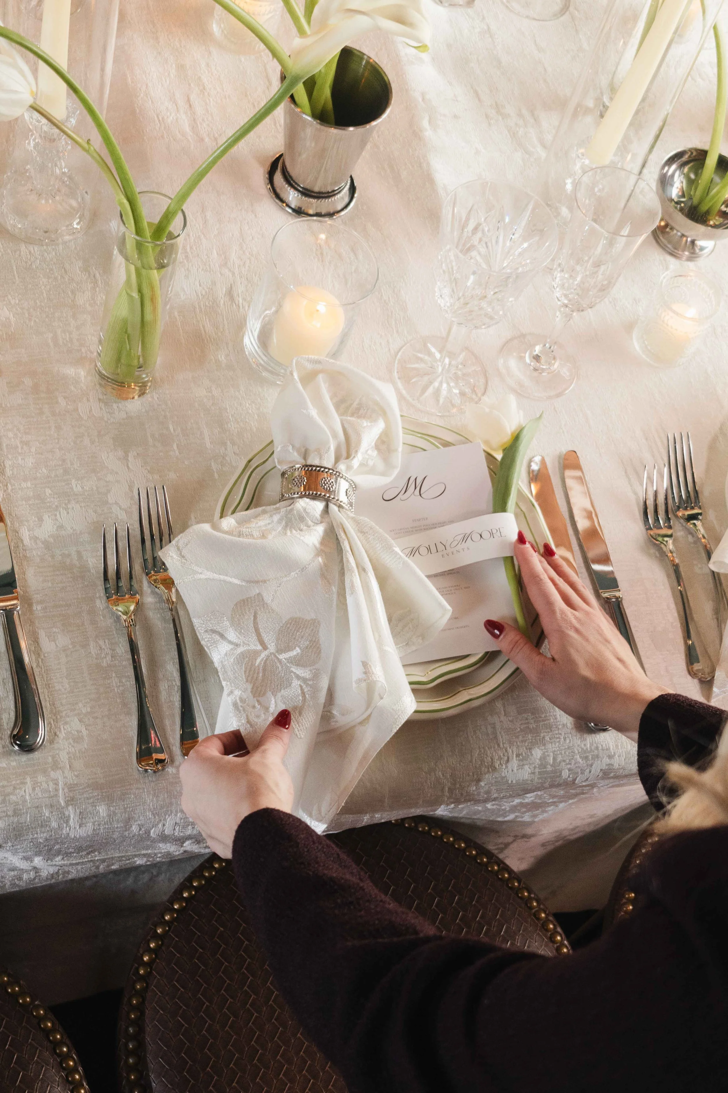 A formal dining table with white tablecloth, floral centerpieces, candles, wine glasses, and silverware. Hands are seen adjusting a decorative napkin on a place setting, with a menu card visible. The setting appears elegant and decorative.