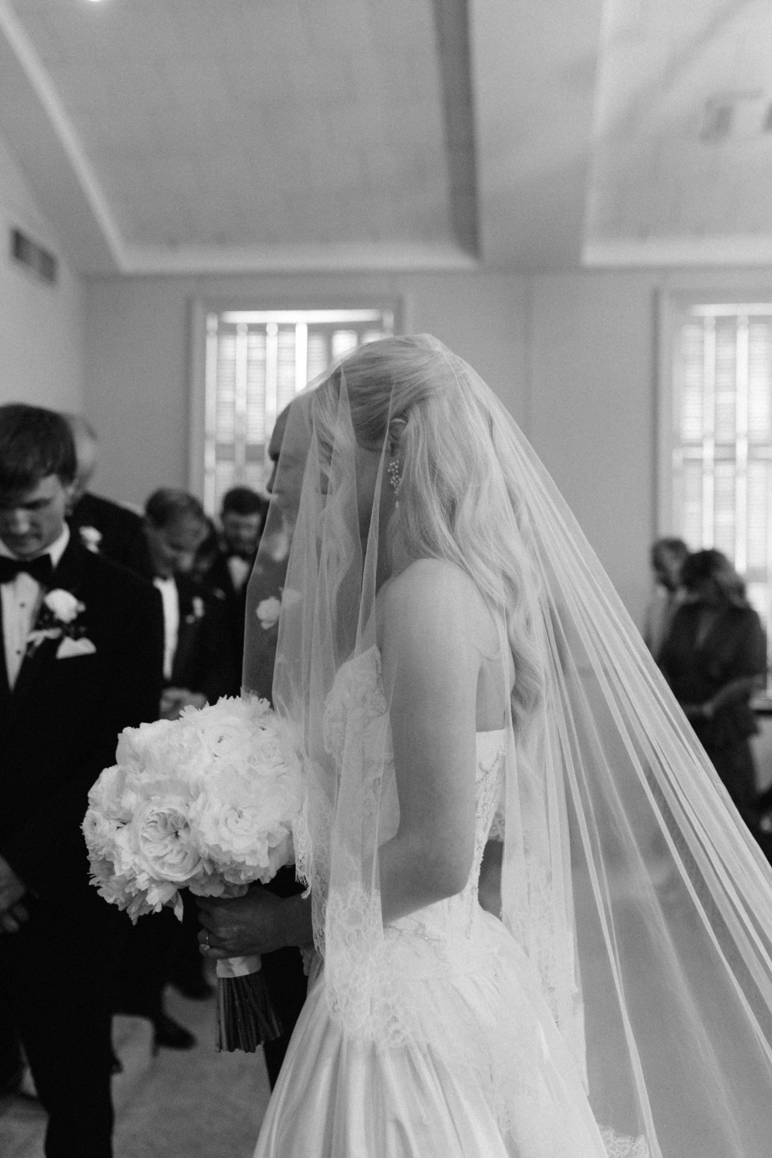 A black and white photo of a bride holding a bouquet of white roses, wearing a wedding dress and veil, during a wedding ceremony inside a room with windows and guests in background.