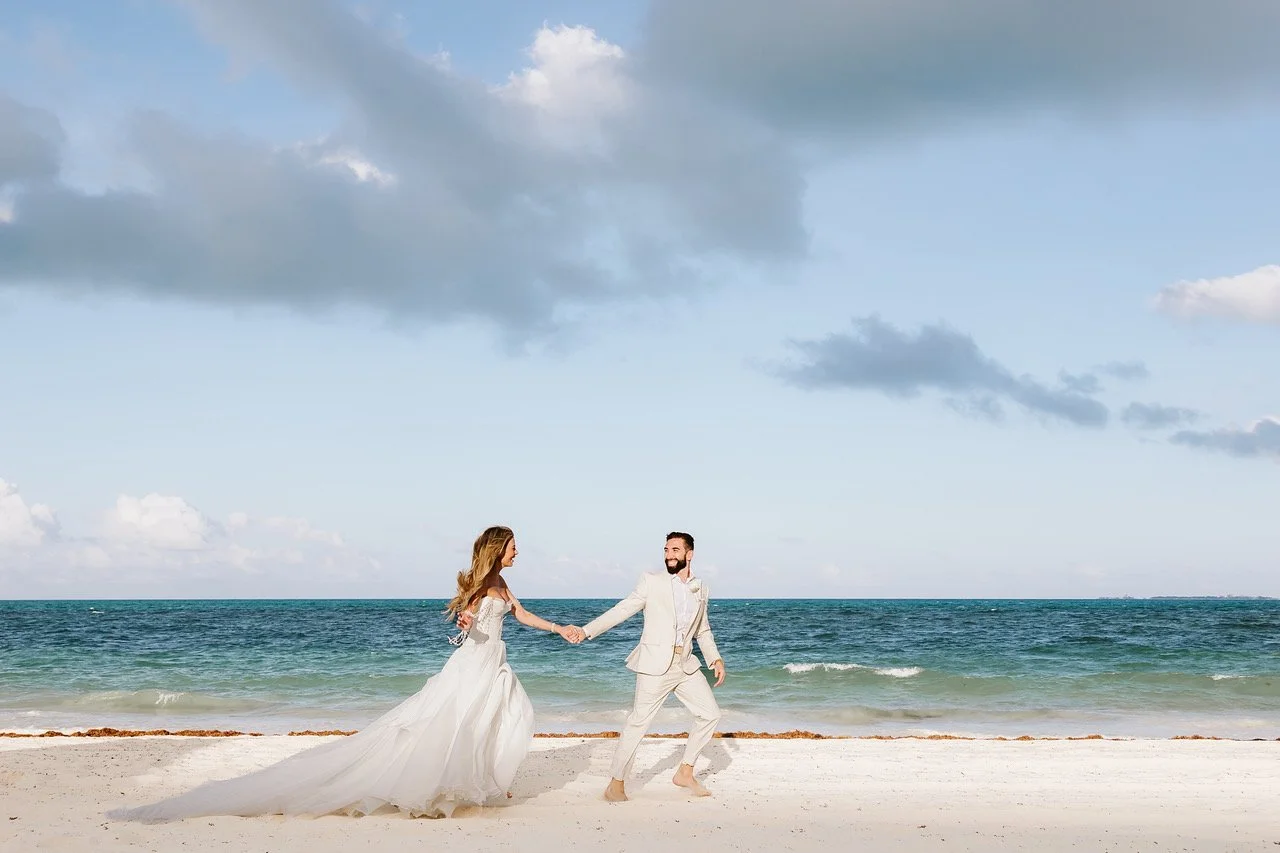 A bride and groom holding hands on a beach, with the ocean and cloudy sky in the background.