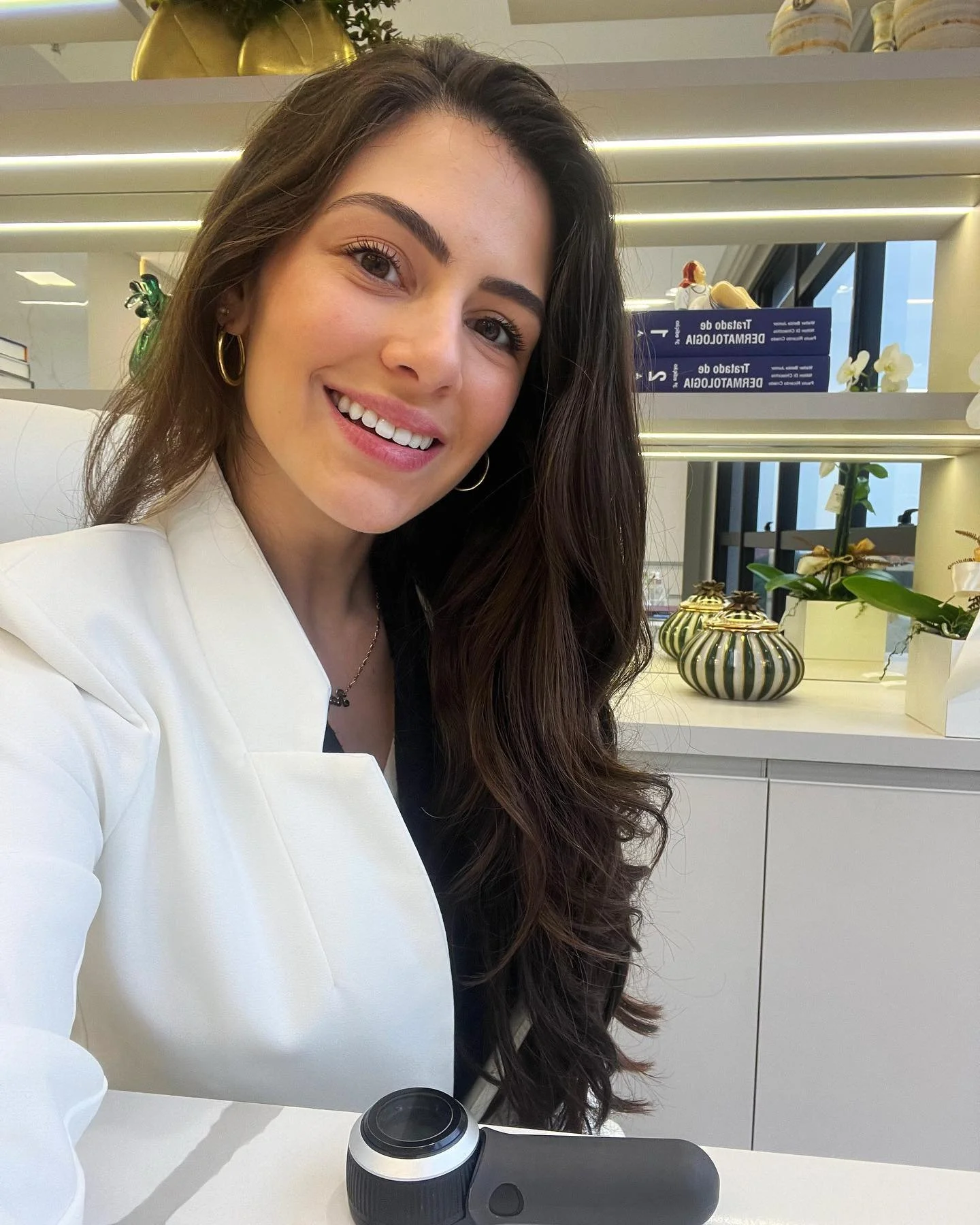 Smiling woman with long dark hair wearing white blazer and gold earrings, sitting at a white table with a camera in front of her, in an indoor space with decorative items and books behind her.