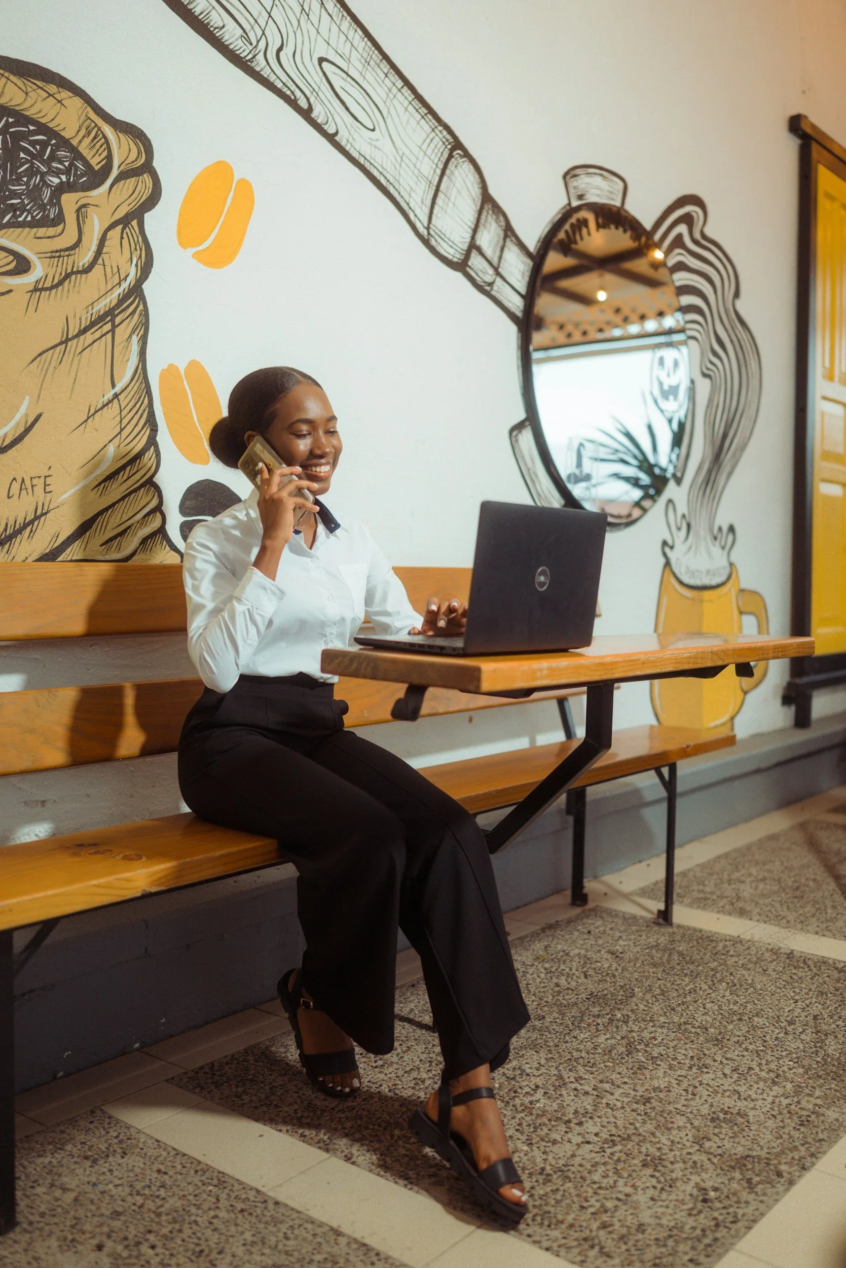 A woman smiling while sitting on a wooden bench in a cafe, talking on her cell phone, with a laptop in front of her, and a colorful wall art mural of coffee beans, yellow leaves, a wooden spoon, and a beer mug in the background.