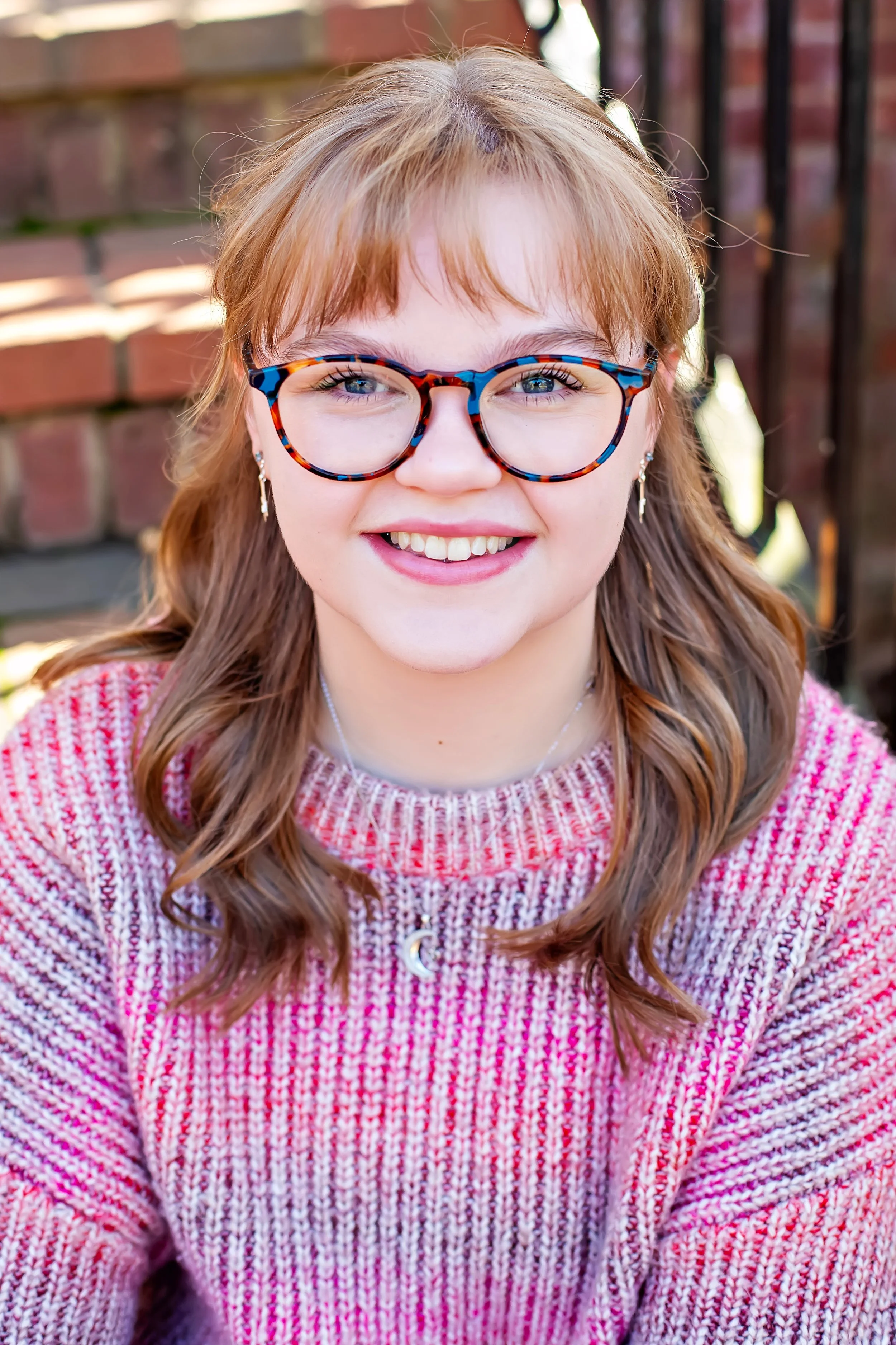 A young girl in her 20's sitting on brick steps. She is smiling at the camera. She has wavy blonde hair with bangs. Her blue eyes can be seen with teal glasses. She is wearing a chunky knit sweater of pink and magenta, and a crescent moon necklace.
