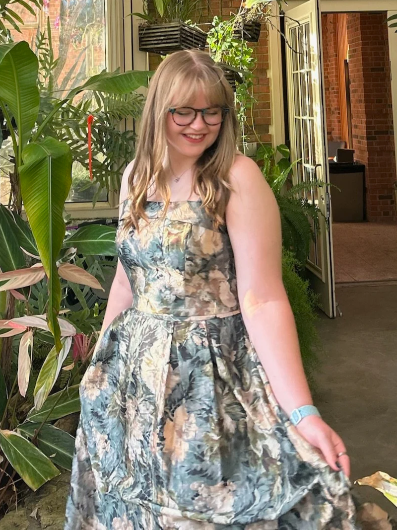 A young woman with blonde hair, glasses, and a floral dress standing indoors among green plants, smiling.