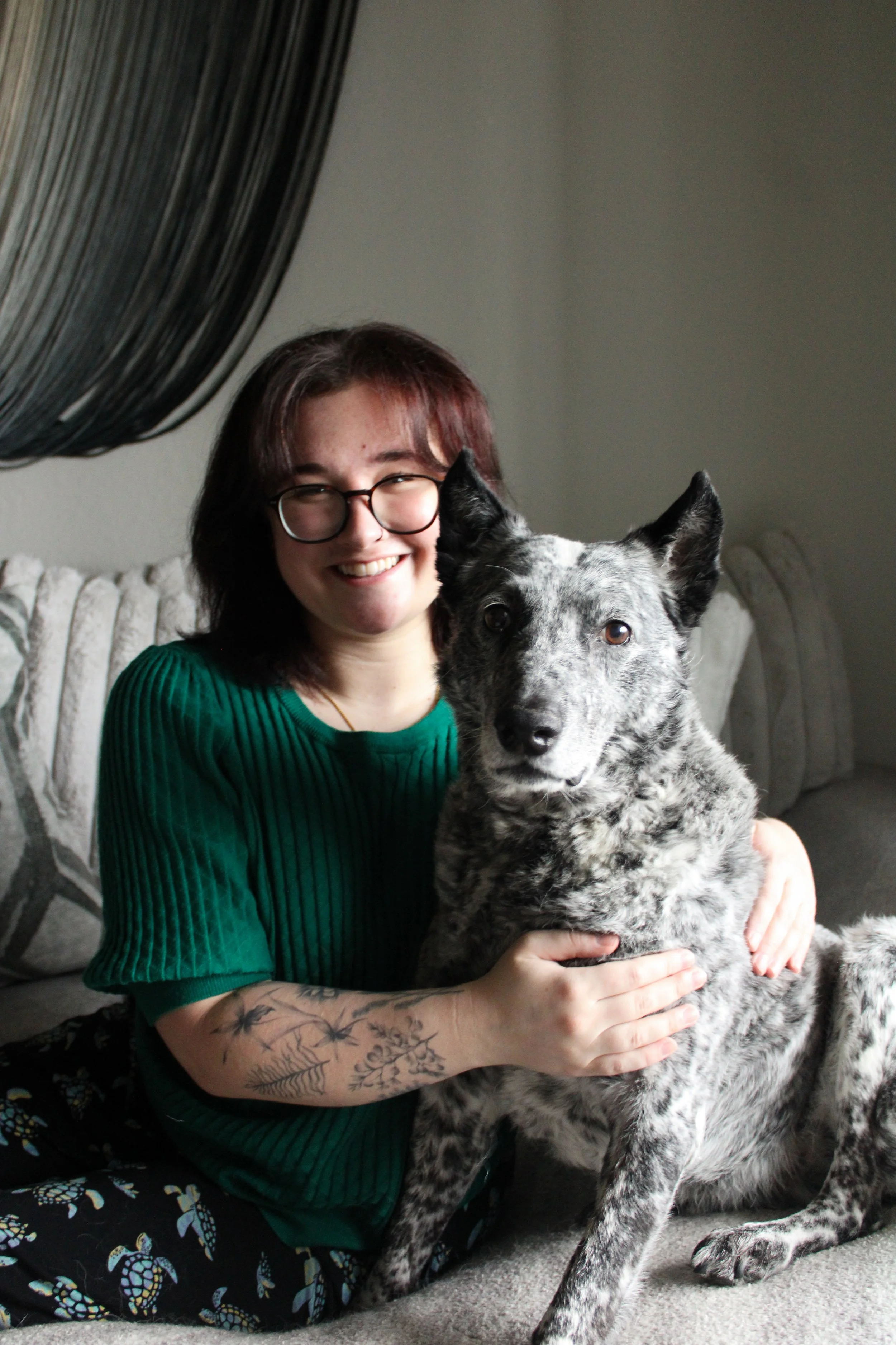 A woman with glasses and tattoos wearing a green top smiles and holds a black and white spotted dog on a gray couch.