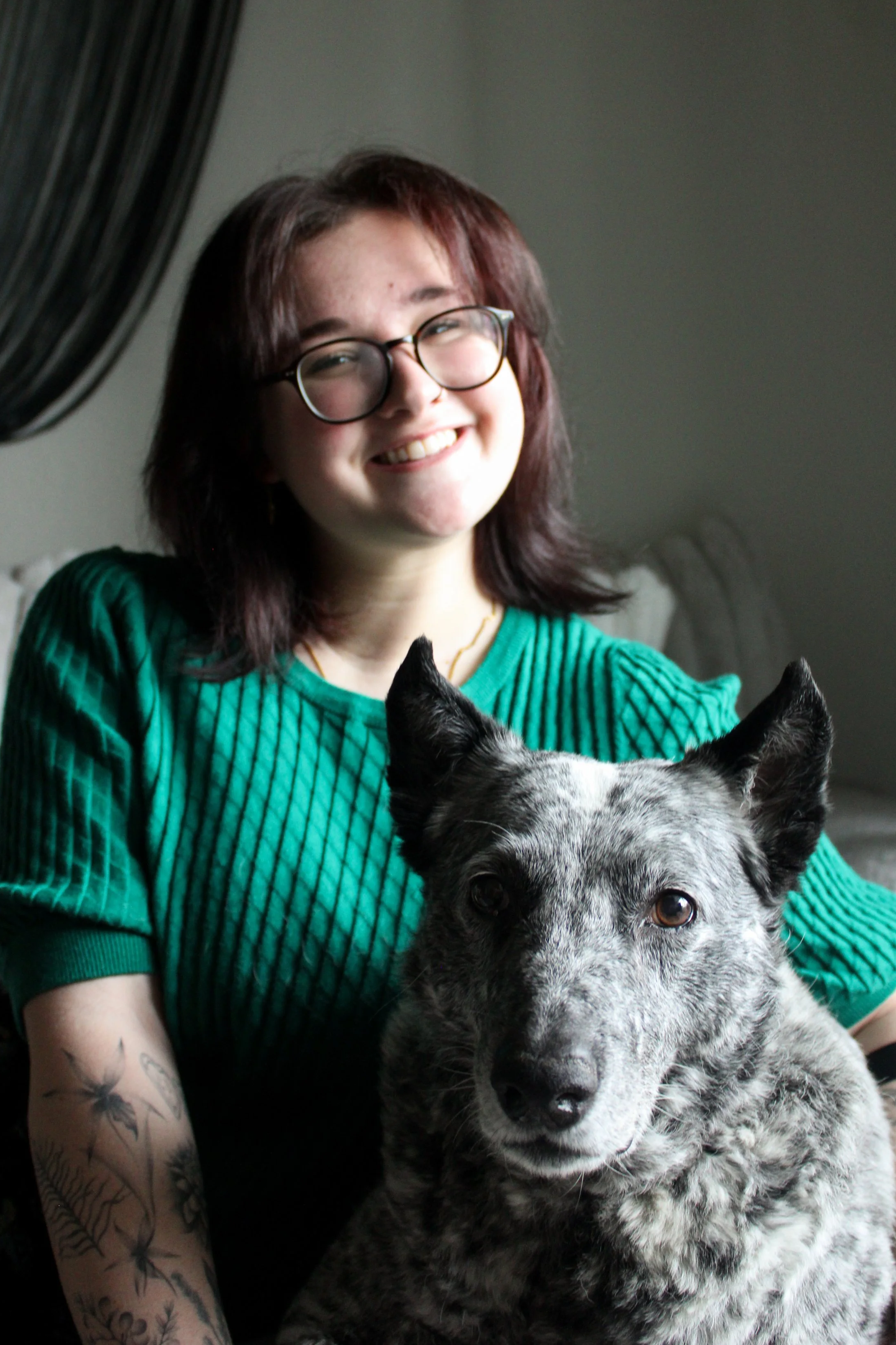 A woman with glasses and tattoos smiling with a speckled black and white dog in front of her.