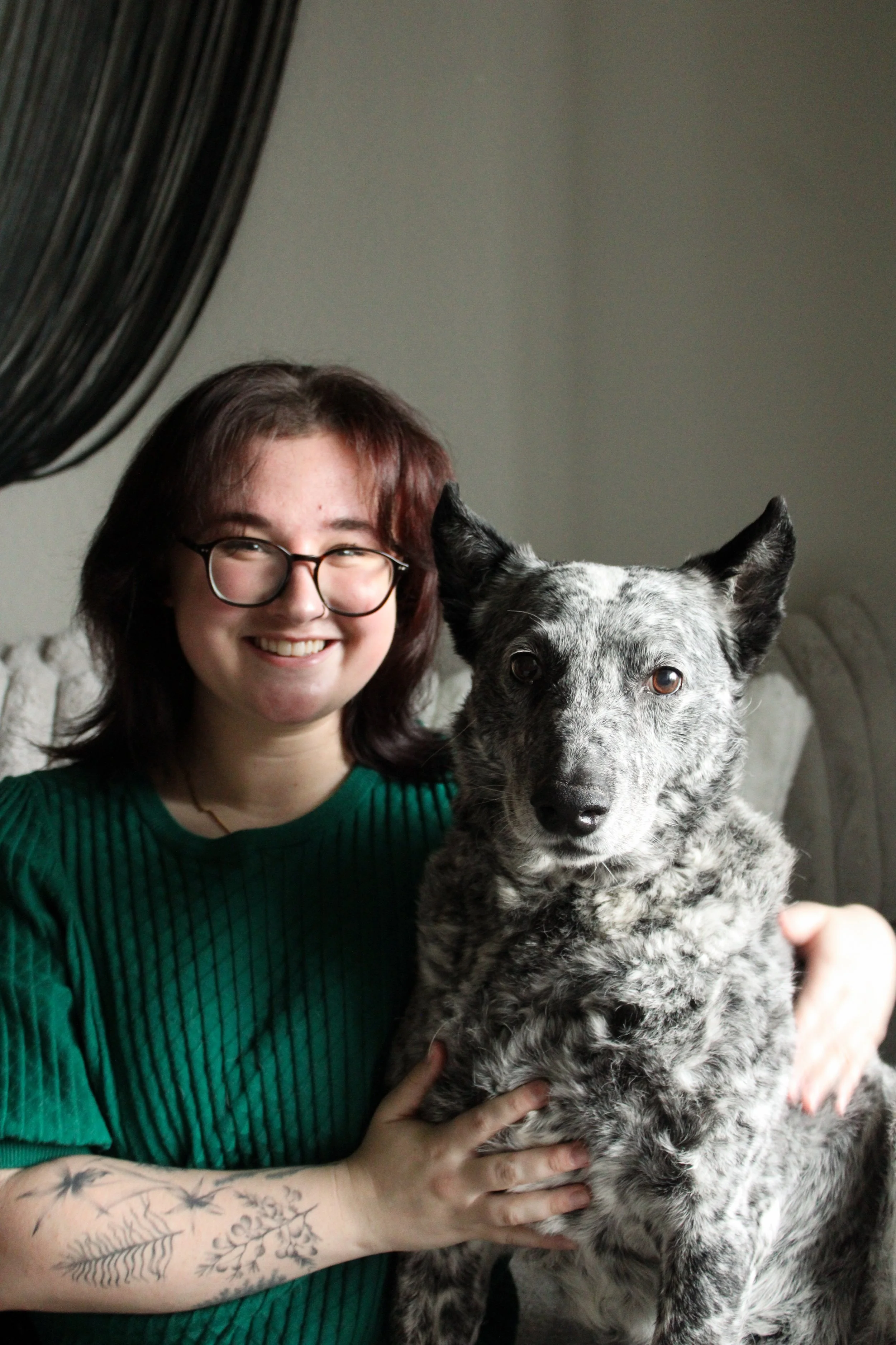 A young woman with glasses and short red hair smiling while holding a speckled black and white dog.