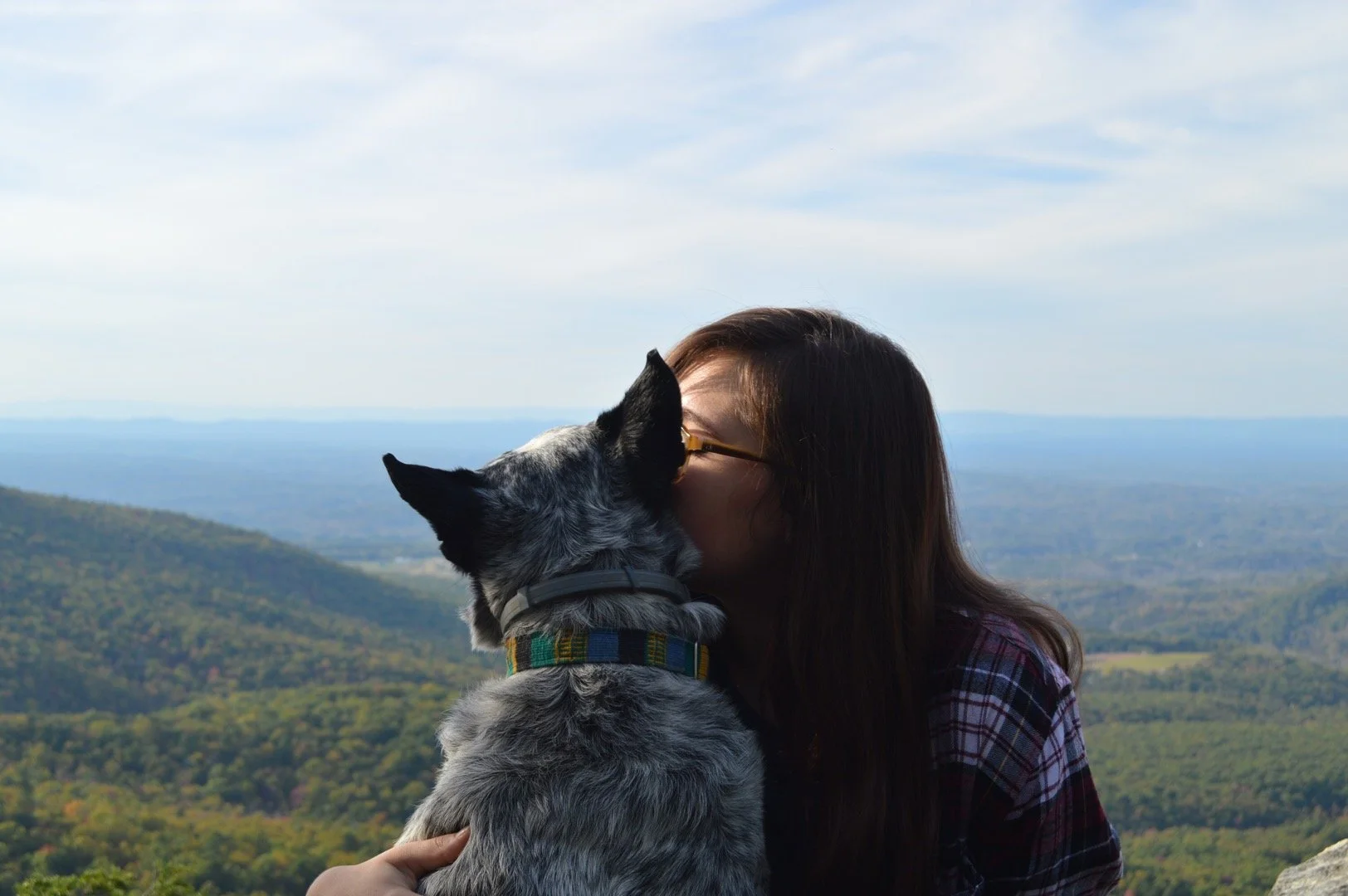 A woman and a dog sharing a kiss against a scenic mountain landscape with a blue sky.