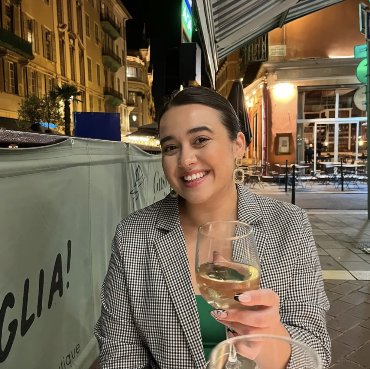 A woman smiling and holding a glass of white wine at an outdoor cafe or restaurant at night, with city buildings and lights in the background.