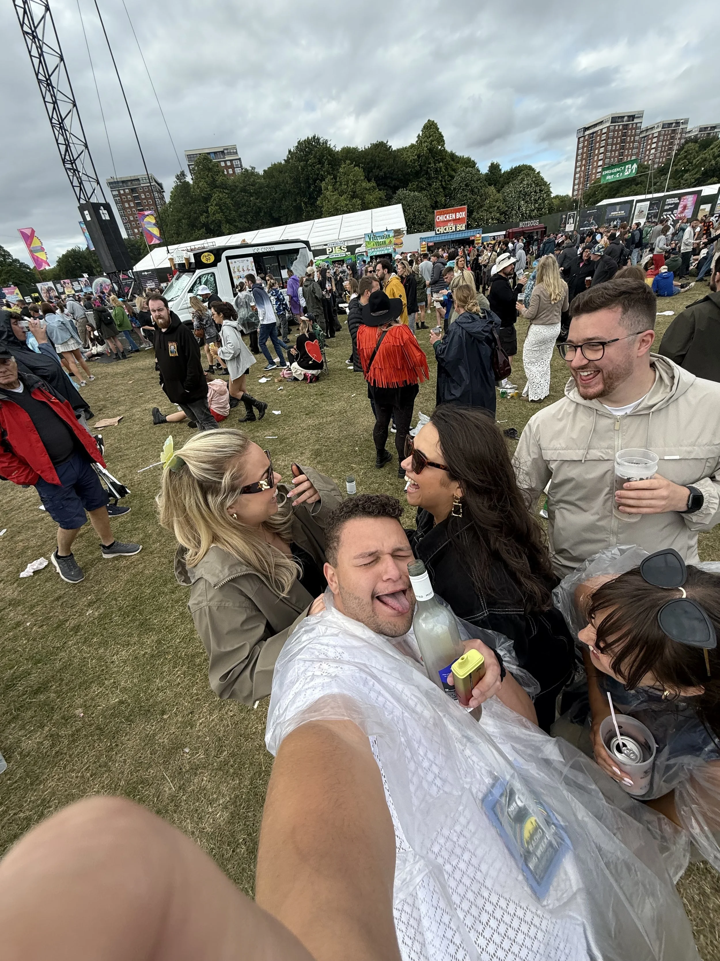 group of friends taking a selfie at an outdoor festival with a large crowd, food trucks, and tents in the background on a cloudy day.