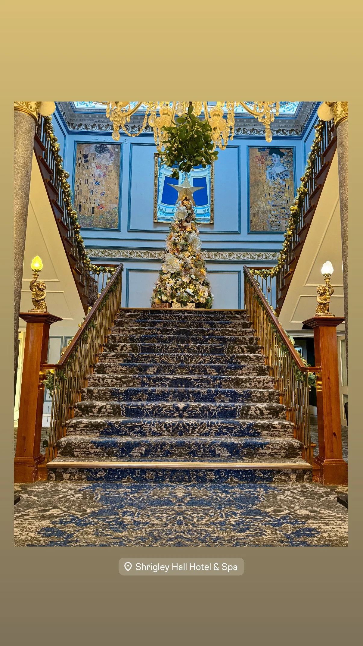 Decorated Christmas tree at the top of a staircase in the Shrigley Hall Hotel & Spa, with ornate blue walls, chandeliers, and artwork in the background.
