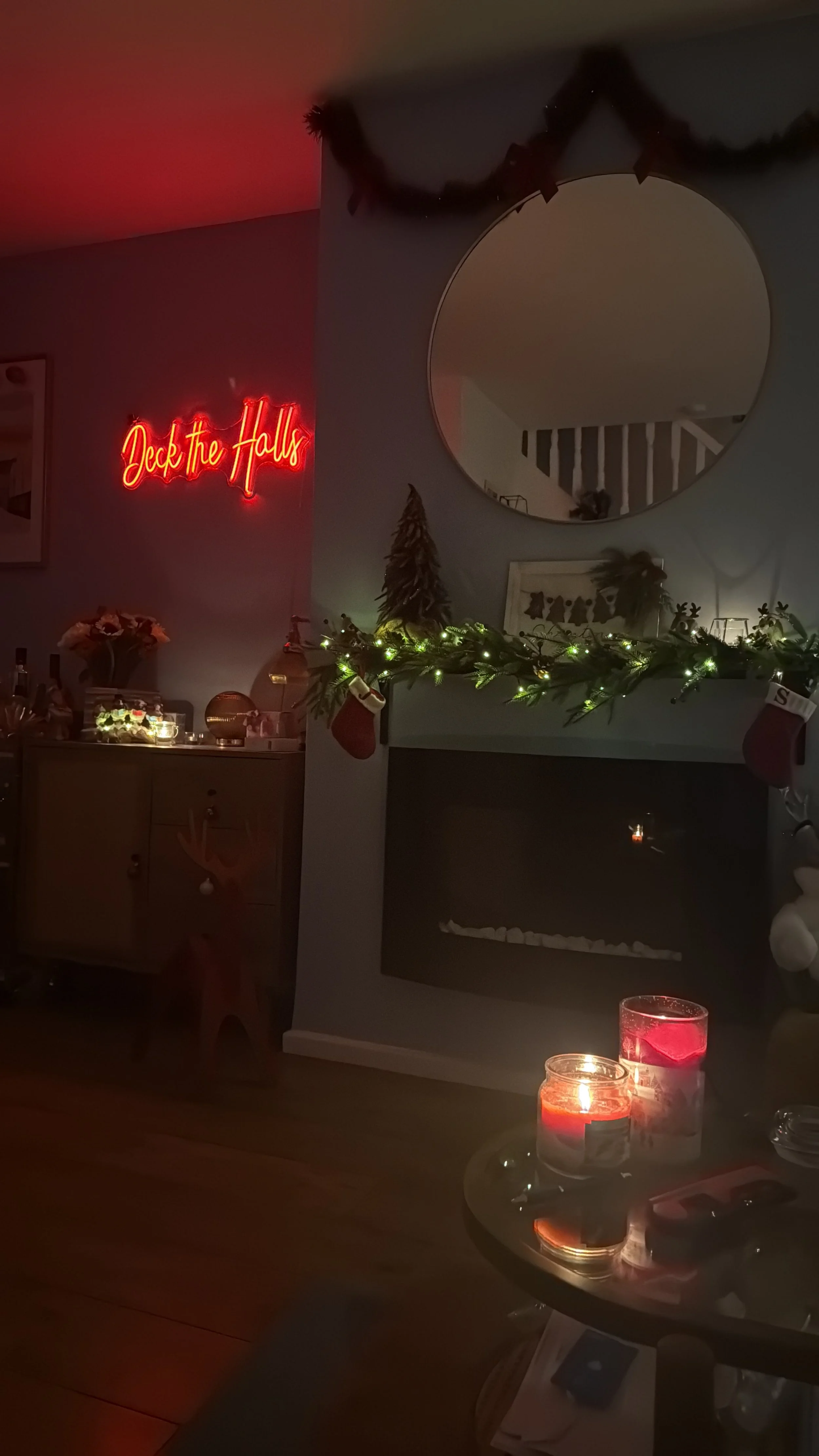 Living room decorated for Christmas with a garland on the fireplace mantel, stockings, a small Christmas tree, and candles. A neon sign reads 'Deck the Halls'.