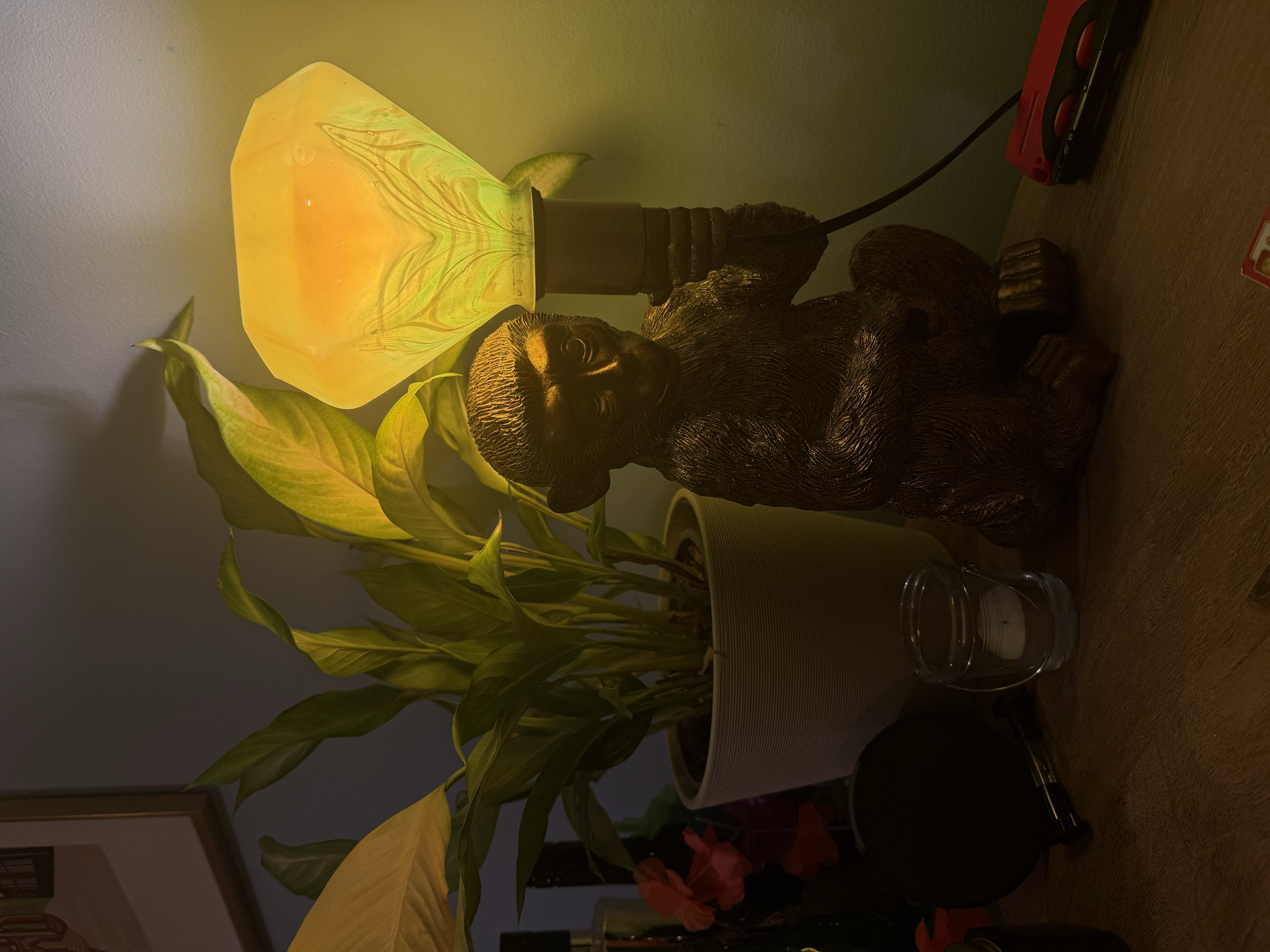 A decorative arrangement on a wooden surface includes a potted plant with large green leaves, a small carved black bear figurine, a yellow floral-shaped lamp, and a clear glass candle holder with a white candle.