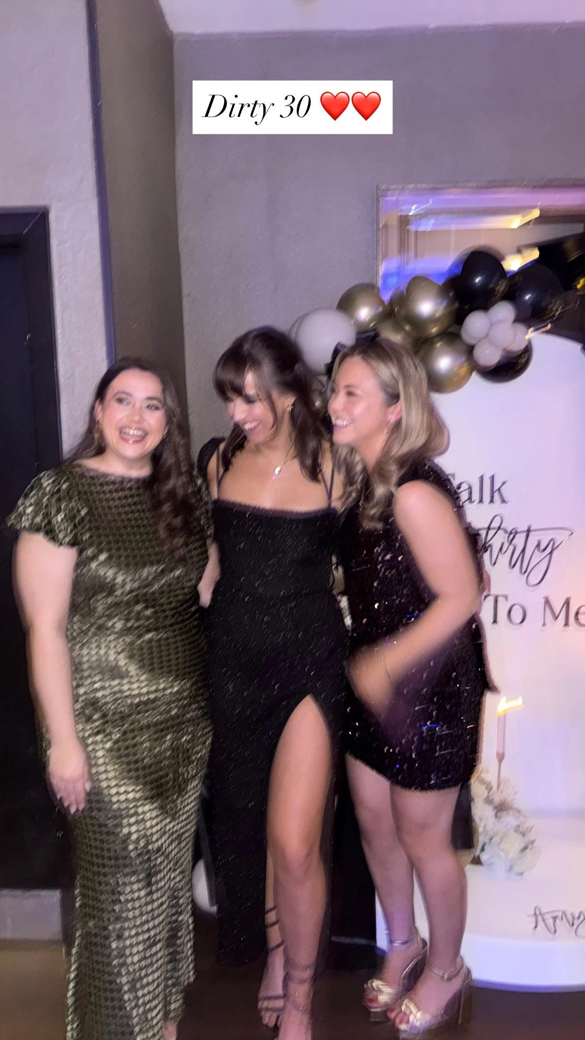 Three women dressed in formal attire at a celebration, standing together and smiling. There are balloons and a sign in the background that says 'Talk Dirty To Me'.