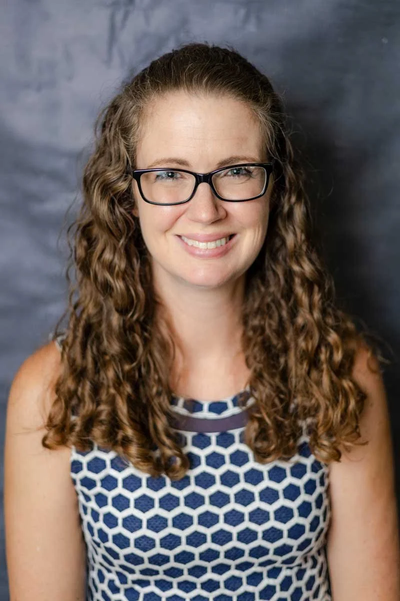 A professor at The Cambridge School of Dallas classical Christian school grades 5-12. A woman with curly brown hair, wearing glasses and a sleeveless top with a navy and white hexagon pattern, smiling at the camera against a gray backdrop.