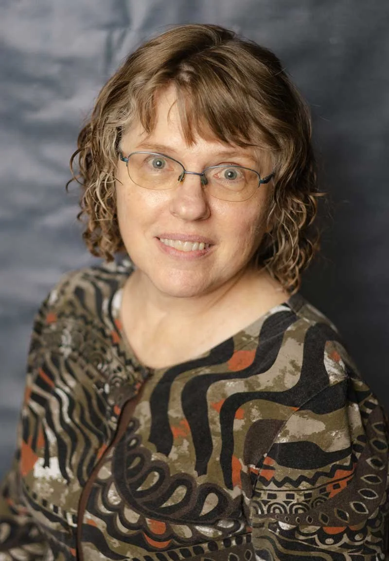 A professor at The Cambridge School of Dallas classical Christian school grades 5-12. A woman with curly brown hair, glasses, and a patterned shirt, smiling in front of a dark patterned backdrop.