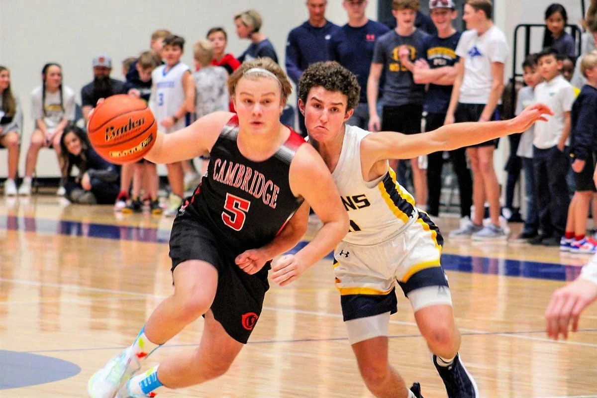 A girl in a black and red basketball uniform dribbling a basketball as she drives towards the hoop, with a player in a white and yellow uniform defending her on a basketball court, other players and spectators watching in the background.