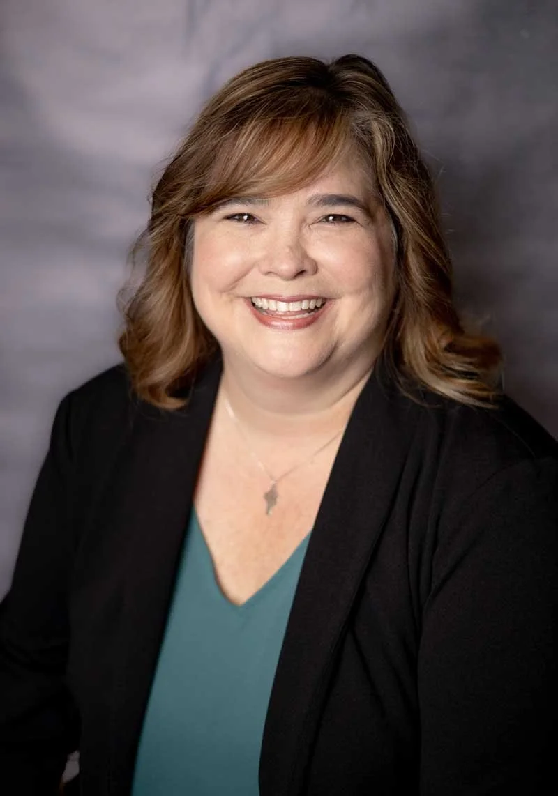 A professor at The Cambridge School of Dallas classical Christian school grades 5-12. A smiling woman with shoulder-length light brown hair, wearing a black blazer over a teal blouse, with a silver necklace, against a blurred gray background.