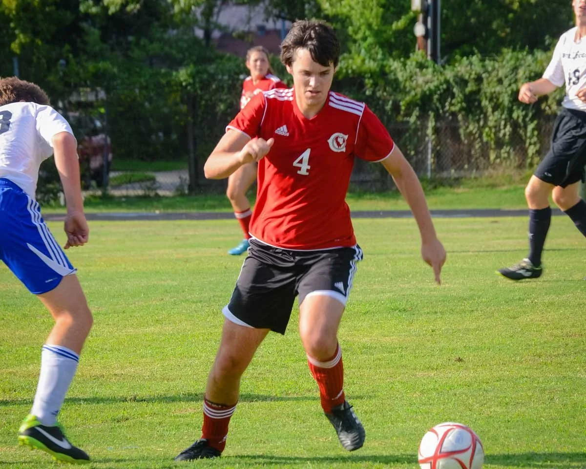A soccer player in a red jersey with the number 4 dribbling a white and red soccer ball on a green field during a game.