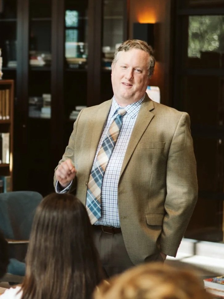 A middle-aged man in a tan blazer, plaid shirt, and plaid tie standing and speaking to a group of people in a room with wooden bookshelves and large windows.