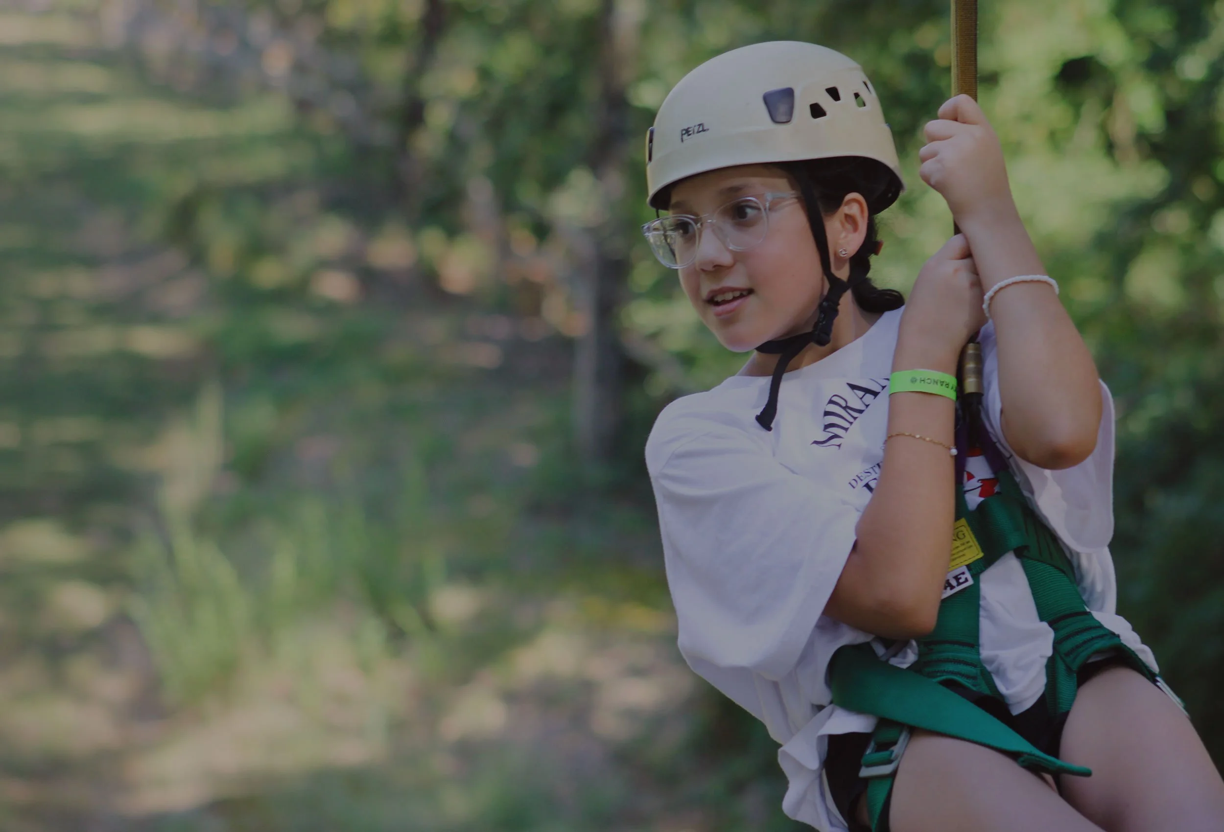 Young girl wearing a helmet and harness, holding onto a rope while ziplining through a forest.