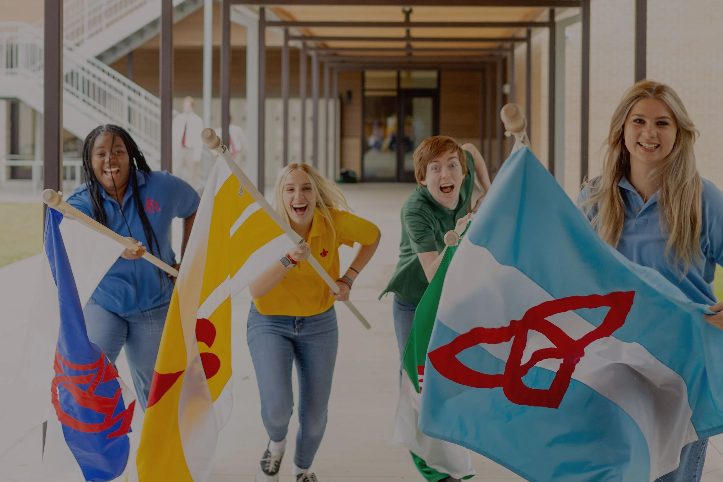 Four young people, two girls and two boys, happily running outdoors while holding colorful flags with symbols and designs, with a school building in the background.