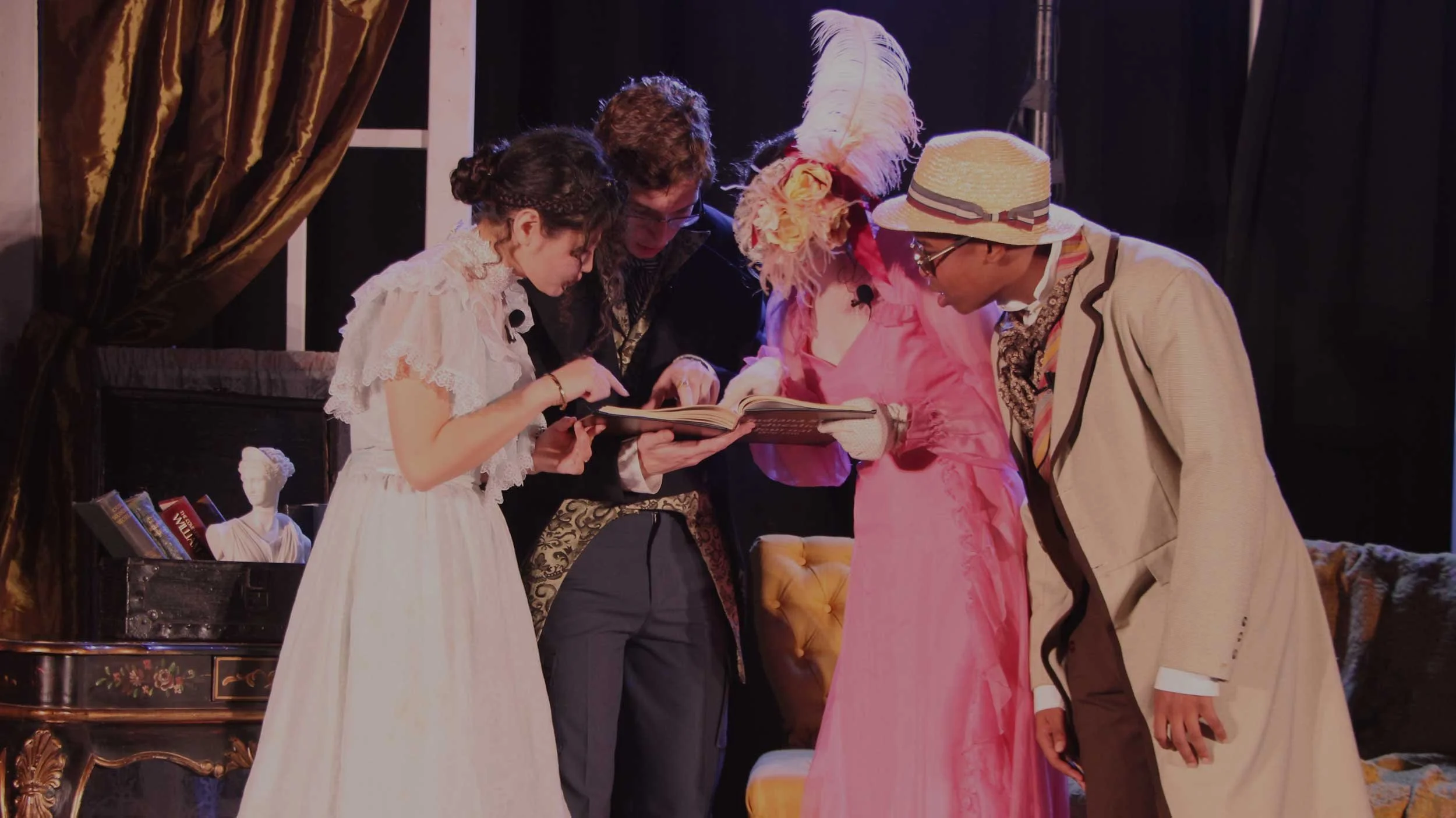 Four people dressed in vintage clothing gathered around and looking at a book on stage decorated with books and a bust of a woman.