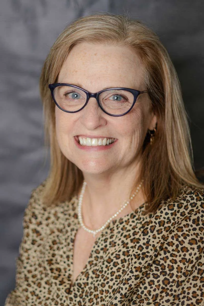 A middle-aged woman with blonde hair, glasses, and a leopard print blouse smiling at the camera. She is wearing a pearl necklace and earrings, with a gray background.