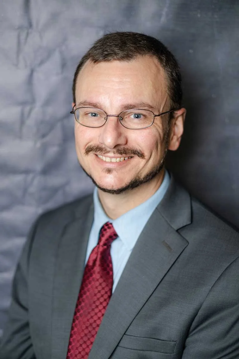 A professor at The Cambridge School of Dallas classical Christian school grades 5-12. A man in a gray suit, light blue shirt, and red patterned tie, smiling with glasses, in front of a dark textured background.