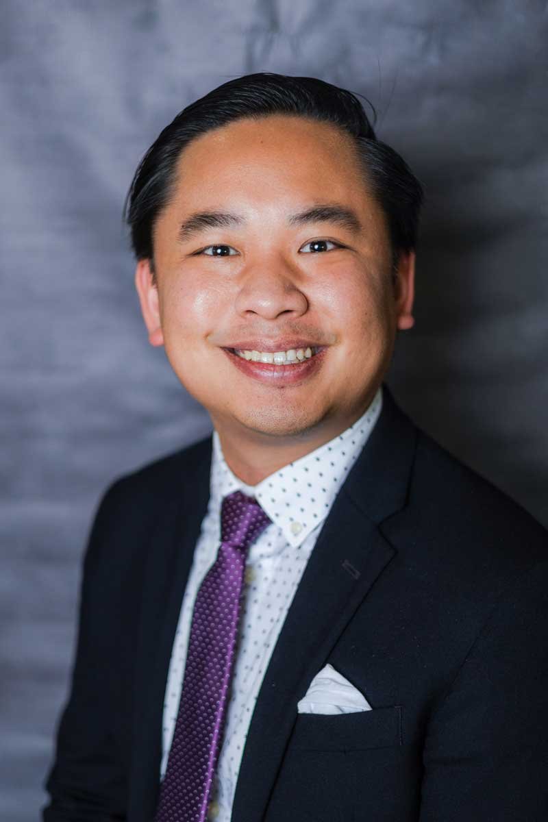 A professor at The Cambridge School of Dallas classical Christian school, smiling with short black hair, wearing a dark suit, white dress shirt with small black dots, a purple tie, and a white pocket square, standing in front of a gray background.