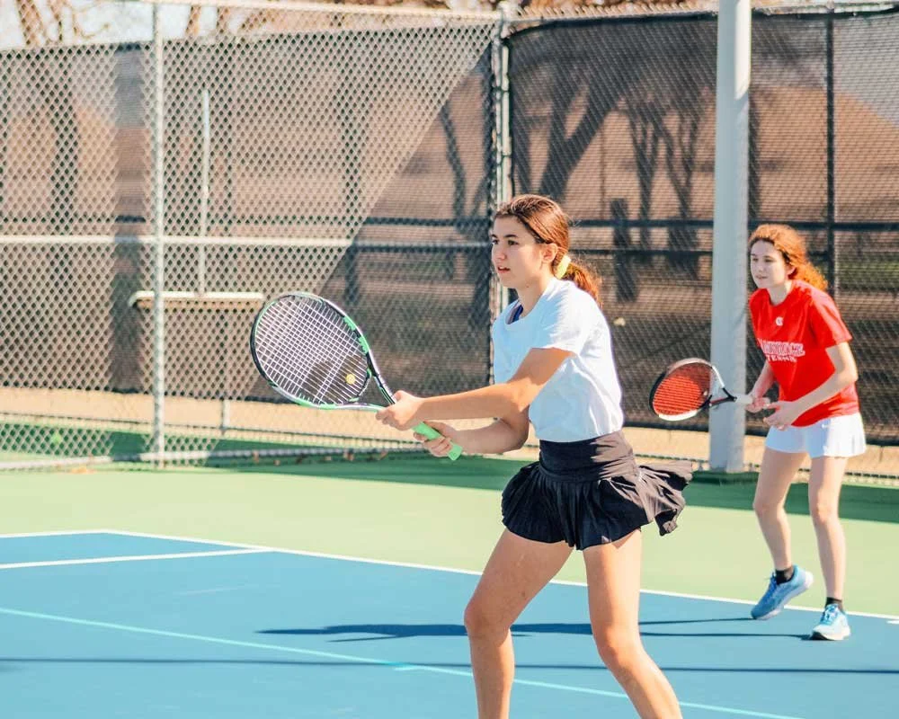 Two young women playing tennis on an outdoor court, with one preparing to hit the ball and the other in position behind her.