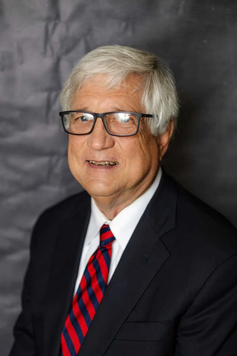 A college counselor at The Cambridge School of Dallas classical Christian school grades 5-12 wearing a dark suit, white shirt, and red striped tie, standing against a dark gray background.