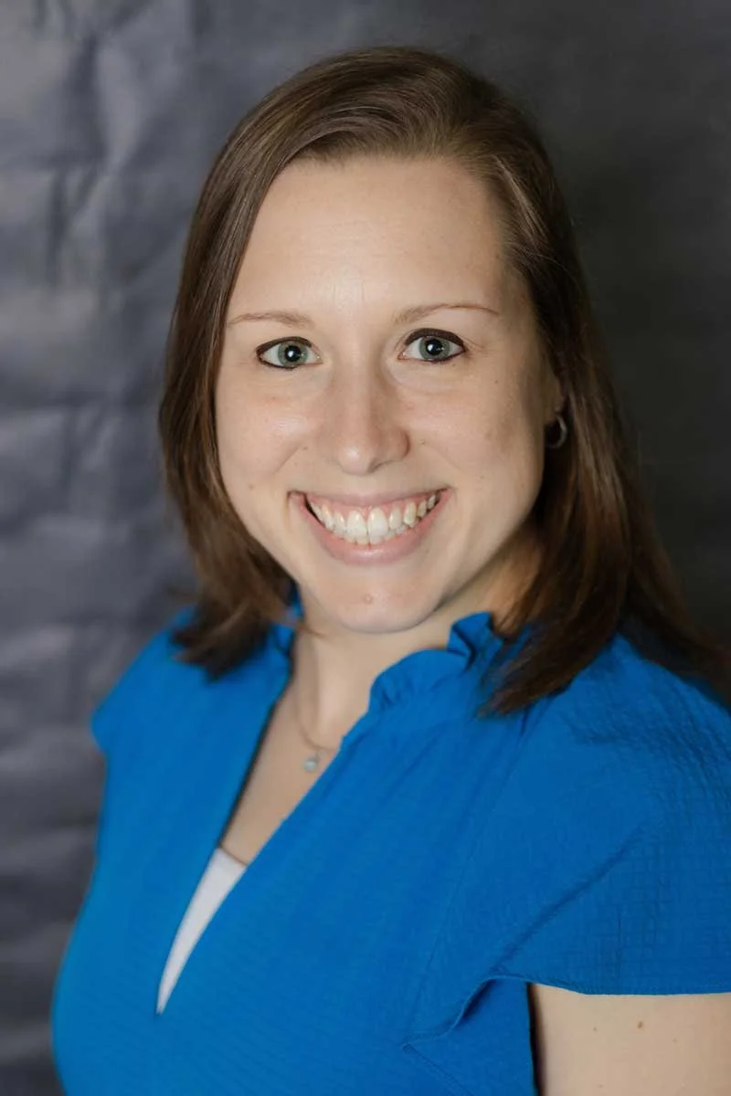 A professor at The Cambridge School of Dallas classical Christian school grades 5-12. Portrait of a woman with shoulder-length brown hair, wearing a blue blouse, smiling against a dark grey background.